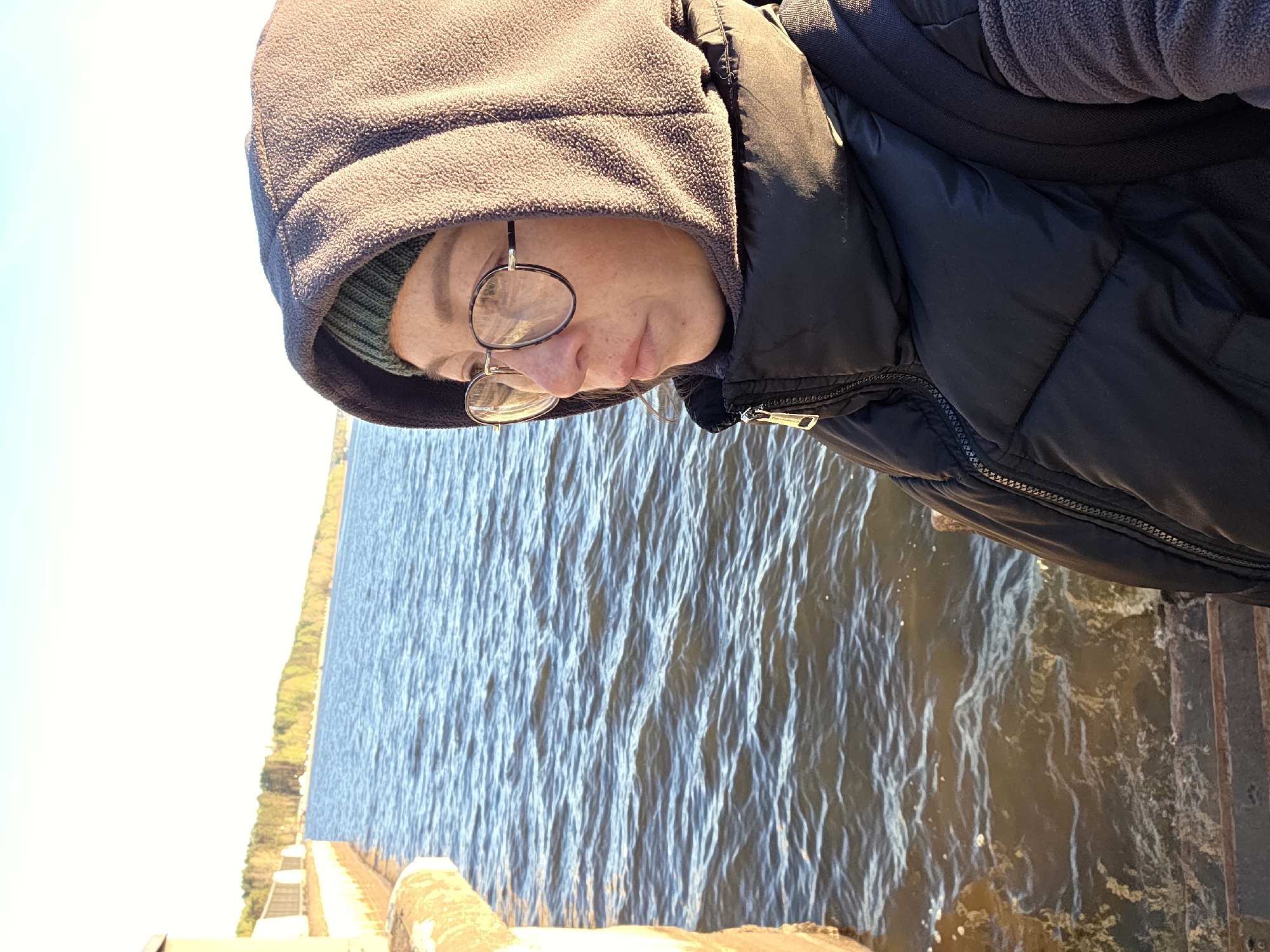 A person in a hooded jacket and glasses stands near the edge of a body of water, possibly a lake or a river, as indicated by the presence of waves and the visible shoreline. The person is wearing a dark-colored puffer jacket over a fleece garment, and the hood is pulled up, revealing a glimpse of a green beanie underneath. They appear to be looking down at the water. The location seems to be a pier or a walkway extending over the water, with visible steps and metal railings. The image suggests an outdoor setting during daytime, potentially with cooler weather, given the heavy clothing. The exact location within Chaikovsky, Russia is not discernible from the image alone.