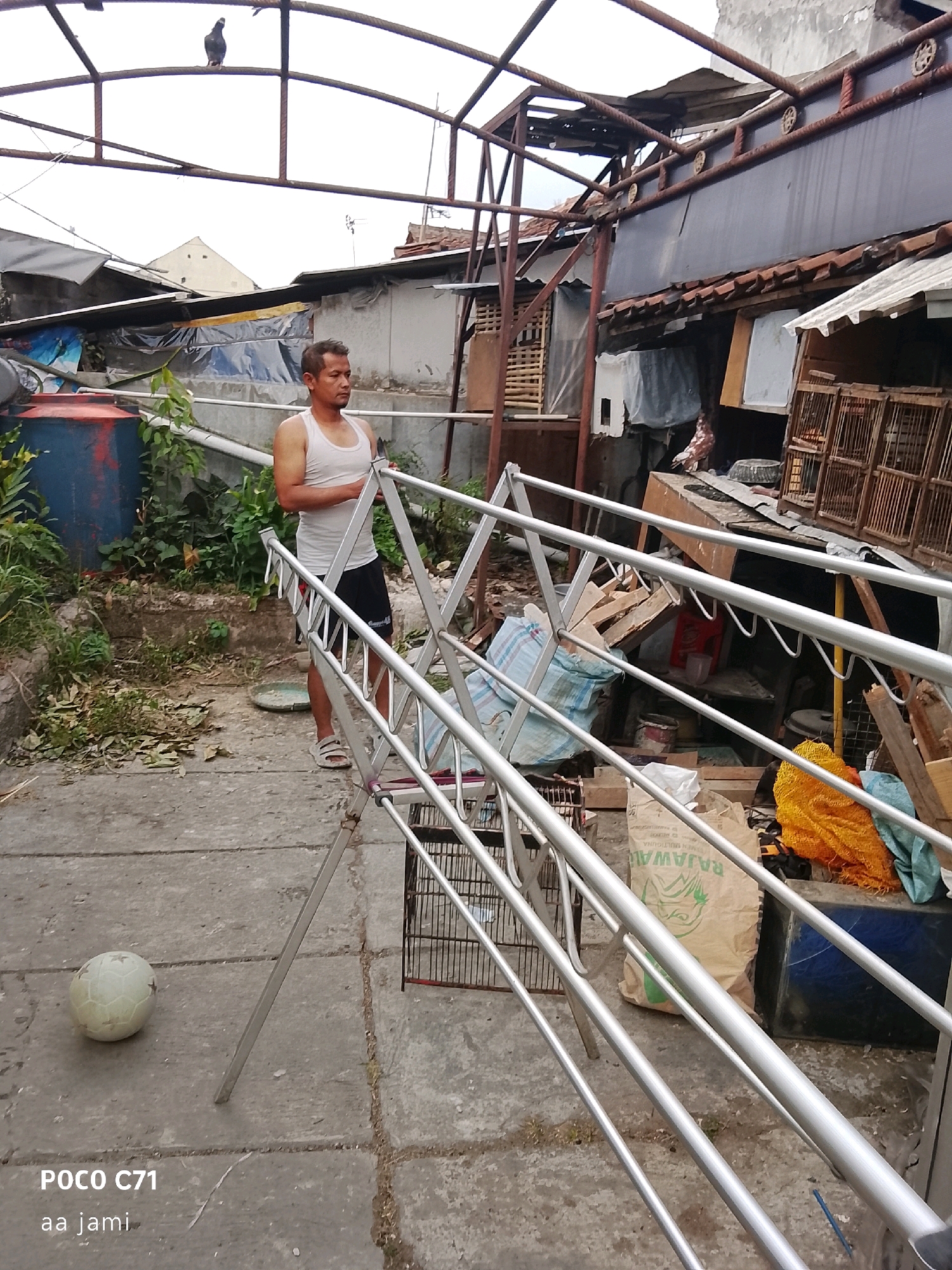 In Tasikmalaya, Indonesia, a man in a white tank top and black shorts stands next to a metal drying rack. He is looking to his right. In the foreground, a white soccer ball lies on a paved surface. The background shows a cluttered yard with various objects including a blue water tank, plants, several bird cages, sacks of material, and wooden structures. A bird is perched on a metal beam overhead. The scene appears to be an outdoor residential area.