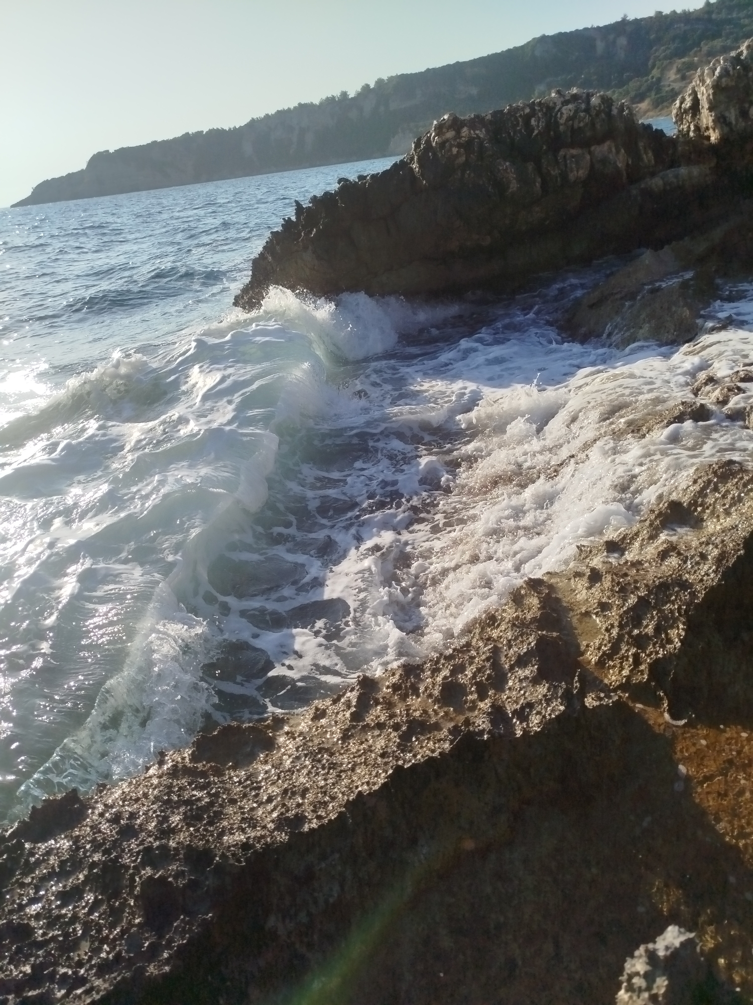 A close-up, low-angle shot captures a crashing wave against rugged, sun-drenched rocks on the coast of Karaburun, Turkey. The water is frothy and white as it breaks, with sunlight glinting off the surface, creating a dynamic and energetic scene. The rocks, in the foreground and mid-ground, are textured and appear to be porous, with varying shades of brown and gray. In the background, the calm, blue sea stretches towards a distant, tree-covered hillside under a clear, pale sky. No people or animals are visible in the frame.