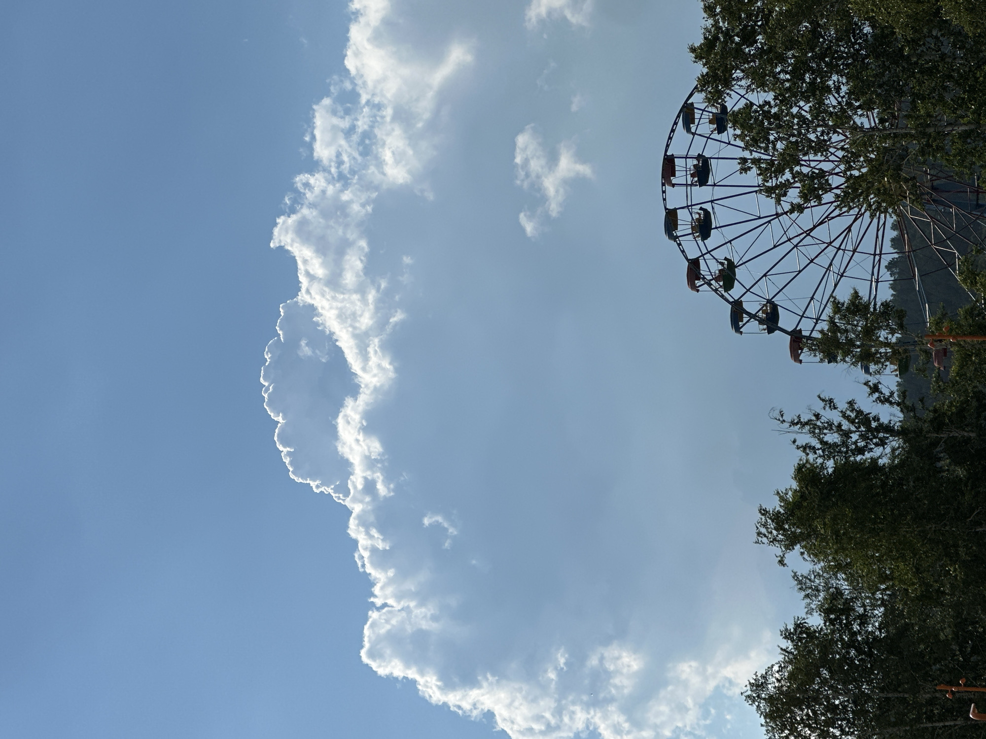 A ferris wheel is partially visible against a bright blue sky with large white clouds. The ferris wheel is dark in color with red and yellow gondolas. Trees line the bottom of the image, partially obscuring the base of the ferris wheel. The lighting suggests it is daytime, likely sunny.