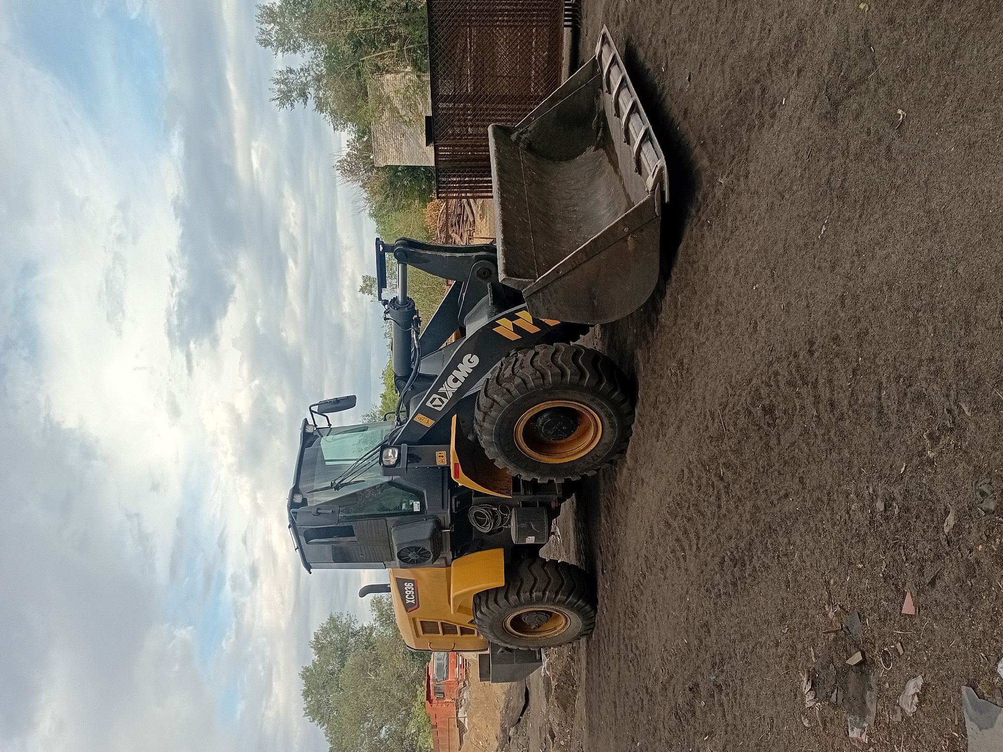 A yellow and black wheel loader, identified as an XCMG XC936, is parked on a dirt lot in Semey, Kazakhstan. The loader is facing towards the viewer's left, with its large bucket lowered to the ground. The machine is in good condition, with no visible damage or rust. The background of the image includes trees, a brick building, and a chain-link fence. The sky is cloudy, with patches of blue visible. There are no people in the image. The lot appears to be a construction site or a storage yard for heavy equipment. The ground is covered in loose dirt and debris, with tire tracks from the loader visible in the soil.