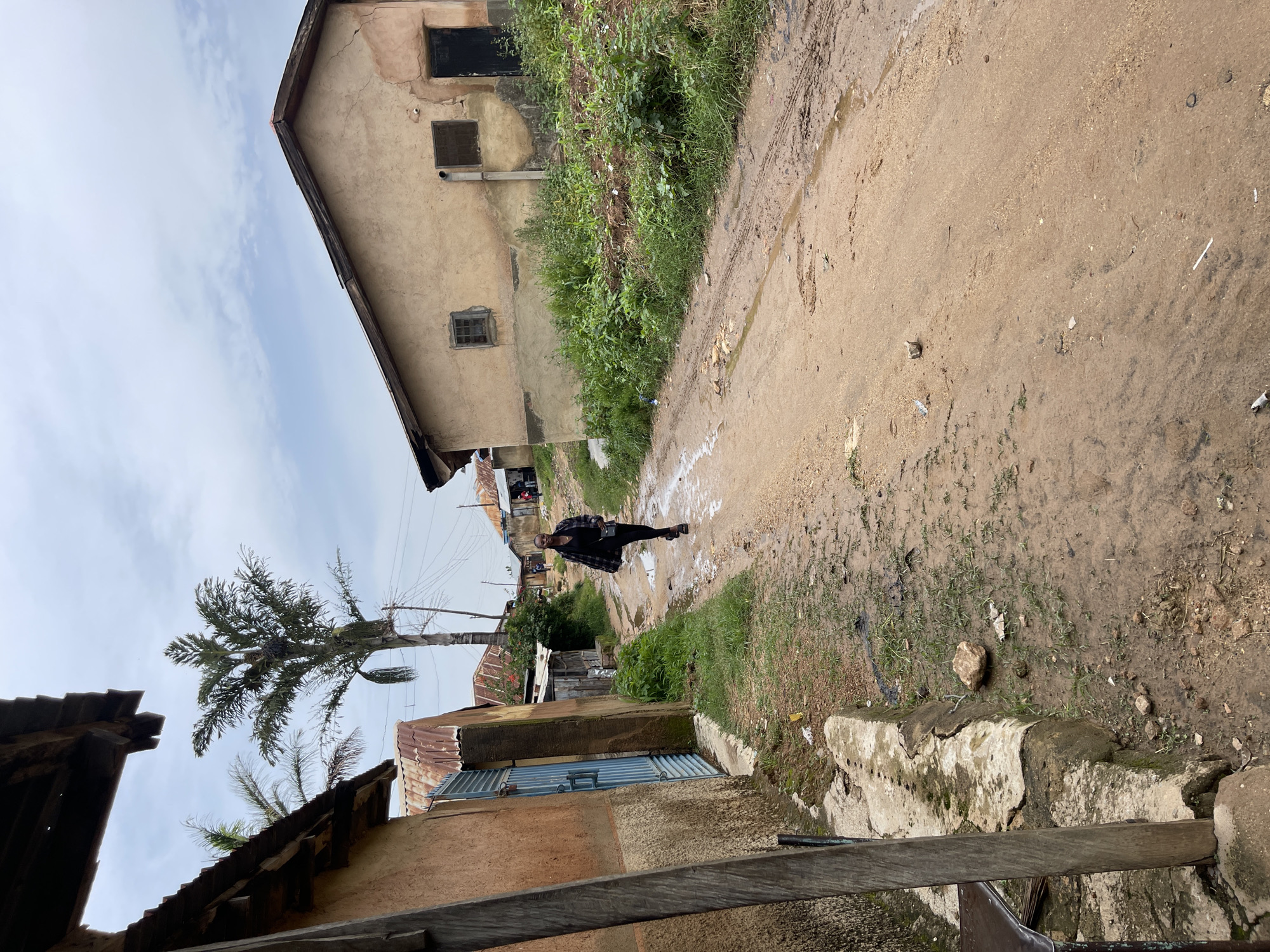 A young Black woman wearing a plaid jacket over a black top and black leggings, and black heeled shoes, walks along a dirt road. She is carrying a black purse on her right shoulder. The road is muddy and appears to have been recently rained on, with puddles and tire tracks visible. To the left of the road are several buildings made of mud and corrugated metal. A tall palm tree stands behind these buildings. To the right of the road are more buildings, one with a tan, textured facade and a dark doorway. The sky is overcast. The overall setting suggests a rural or semi-urban environment in Rafin Par, Nigeria.
