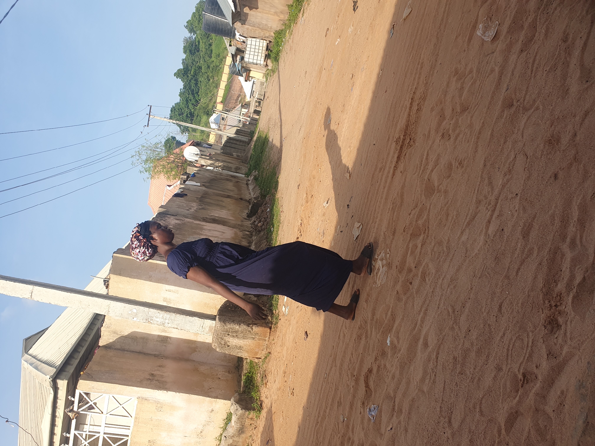 A woman walks on a sandy path in Lokoja, Nigeria. The woman is wearing a navy blue dress and a colorful headscarf. She is walking away from the camera, towards the left side of the frame. Her body is angled to the left, and her right arm is bent at the elbow. The path is made of sand and dirt, and there is some scattered trash on the ground. On the left side of the path, there is a concrete wall and a building with a white roof. On the right side of the path, there are more buildings and vegetation. The sky is clear and blue.