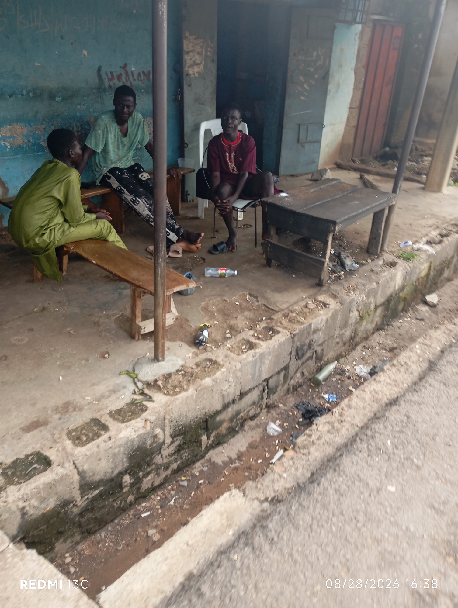 The image depicts an outdoor scene, possibly in the city of Shendam, Nigeria. Three young men are present. One individual, wearing a green traditional outfit, is seated on a wooden bench. Another man, in a green t-shirt and patterned pants, is also on the bench. The third man, wearing a maroon traditional garment, is seated on a white plastic chair. A dark wooden table is positioned in front of him. The environment appears to be a somewhat disheveled area with a concrete floor. A drainage channel runs along the foreground, containing some debris and litter. The background shows a building with blue and brown walls. The overall impression is one of a casual gathering in a functional, perhaps semi-urban, setting.