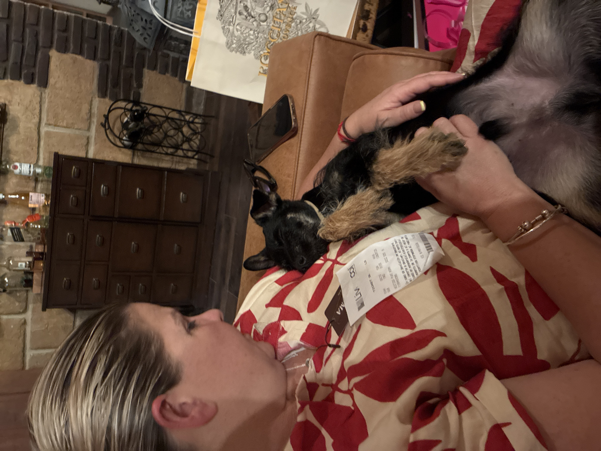 A woman is lying down, holding a small black and tan dog on her stomach. She is wearing a red and white patterned top. The dog is on its back, with its legs in the air. A price tag is visible on the woman's top. A cell phone sits on the side of a brown leather couch. The location appears to be indoors, with a stone wall and dark brown furniture visible in the background. The specific location is Cañada de Madero, Mexico.