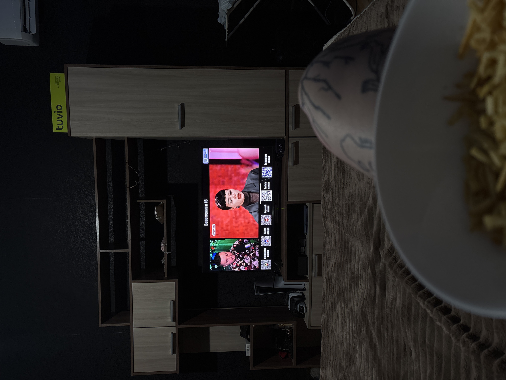 The image shows a television displaying the Russian TV show "Pregnant at 16" (Беременна в 16). There is a TV cabinet with compartments next to the TV. To the right, part of a bowl of what appears to be fries and the arm of a person with a tattoo are visible. The brand "tuvio" is also visible on a label on the TV cabinet. The environment seems to be indoors, and the location in the city of Chornomorskoe, Ukraine cannot be confirmed based solely on the information available in the image.