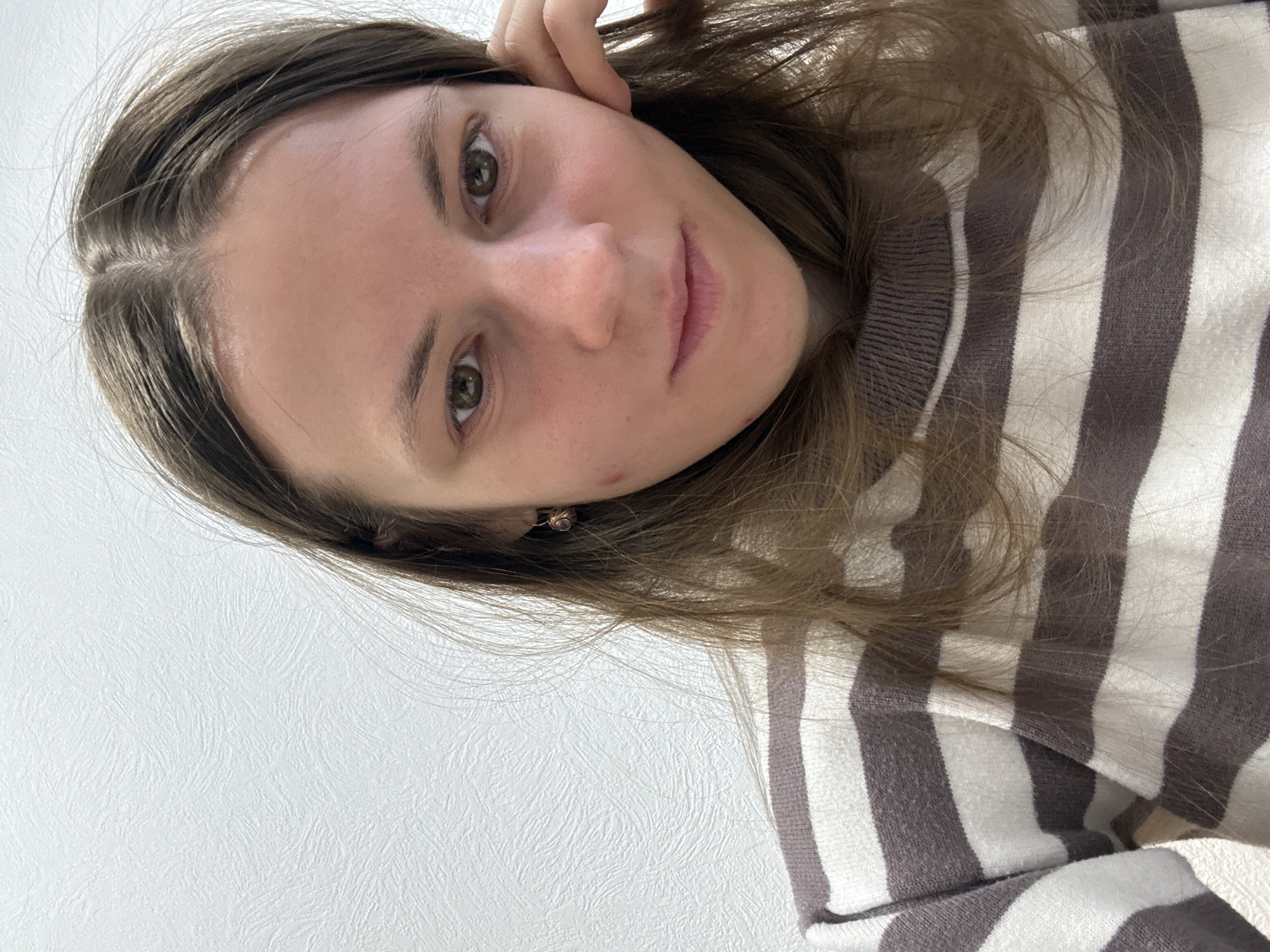 A selfie-style image depicts a young woman with long, light-brown hair wearing a gray and white striped sweater. Her head is tilted towards the right, and she looks directly into the camera. The background is a plain, white surface. There's no indication of the city of Chaikovsky, Russia in the image. No actions or interactions are visible in the image. The environment consists of the plain white background and the presence of the sweater and hair.
