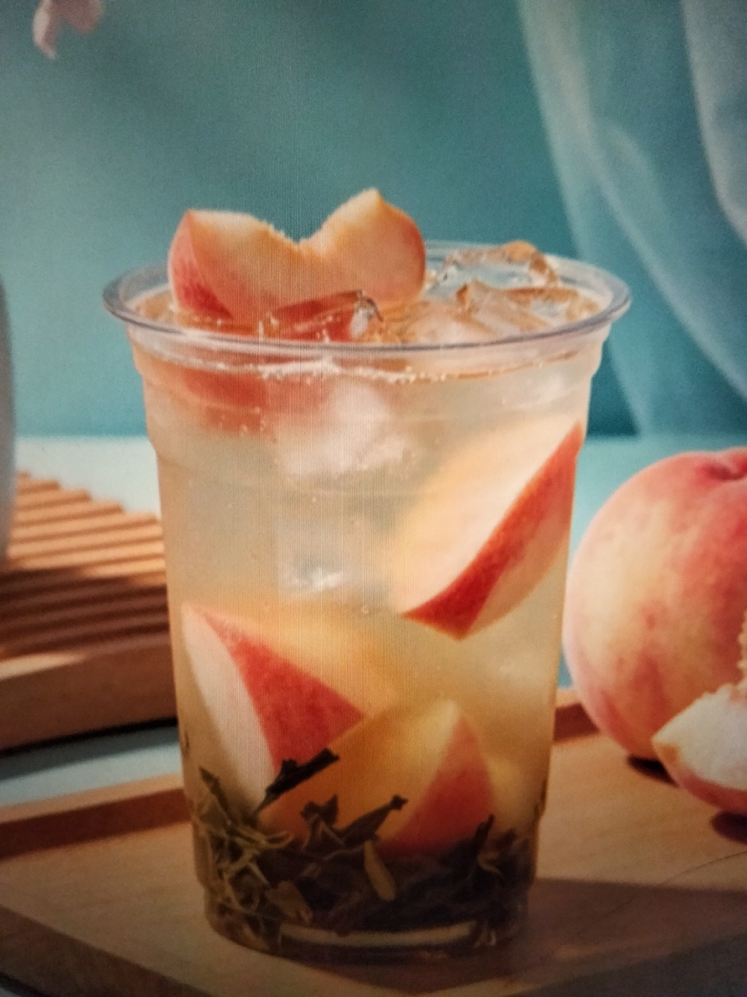 A clear plastic cup filled with a light-colored beverage, ice cubes, and several slices of peach is the central subject. The drink appears to be a refreshing beverage, possibly iced tea or a fruit-infused drink. The cup is decorated with slices of peach, some submerged in the liquid, and others precariously balanced on the rim. In the background, out of focus, is a whole peach and some sliced peaches. The scene is set on a wooden surface, with a blurred background of what appears to be a light blue curtain or fabric. There are no people or discernible actions in the image. The lighting suggests a bright, possibly natural light source, illuminating the drink and fruit. The overall impression is one of a refreshing and visually appealing beverage. The image does not contain any information to suggest its location in Payagaboh, Indonesia.