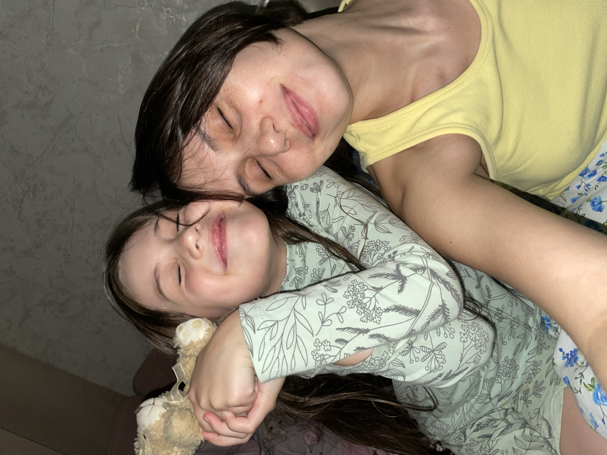 A woman and a child are lying close together, seemingly asleep, in an indoor setting. The woman has dark hair and is wearing a yellow top and floral print bottoms. The child, who also has dark hair, is wearing a green and white patterned long-sleeved top. The child is holding a small, light-colored teddy bear. The setting appears to be a bedroom, based on the bed. There are no indicators of the city of Izhevsk, Russia.
