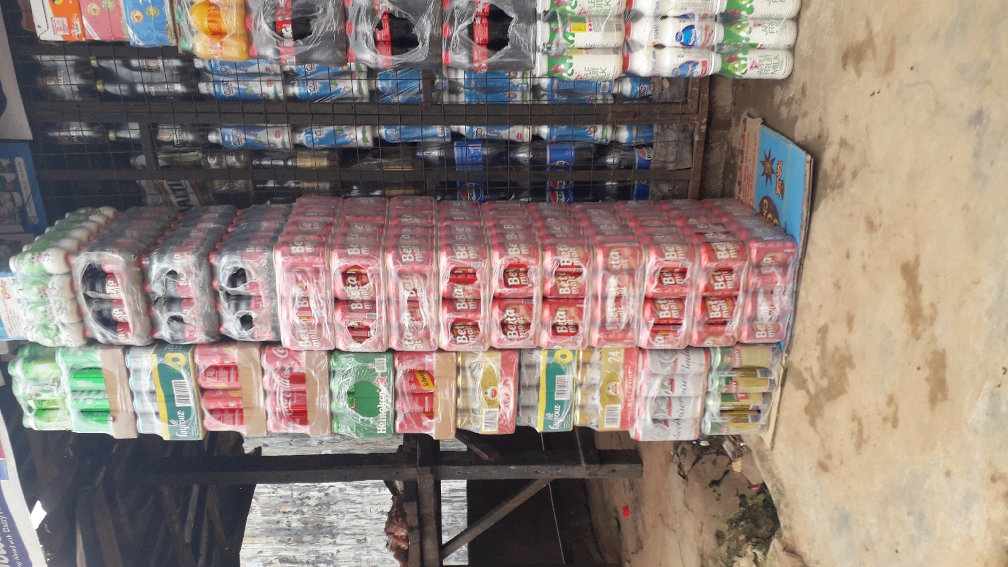 This is a forensic, matter-of-fact description of a street-side beverage stall in Nkpor, Nigeria. The scene is cluttered with an assortment of bottled and canned drinks stacked high on display. The dominant colors are red, green, and white, reflecting the packaging of the various beverages.
In the foreground, the ground appears to be a mix of concrete and dirt, with some damp patches. A large pile of red and white cans, possibly beer or malt drinks, is stacked in the center of the image, shrink-wrapped for protection. To the left of this central stack, more canned drinks in green and yellow packaging are visible, along with some red cans. Further to the left, a metal shelving unit holds various items, including what appears to be raw meat on a tray at the top, and more canned drinks below.
To the right of the central stack, a metal grate fence encloses additional beverage products, including bottles with blue and white labels, and some dark colored bottles that could be soda. Higher up on the right, more packaged beverages are visible, including some with orange and yellow labels. At the very bottom right, a stack of bottles with white and green labels, possibly milk or juice, is arranged.
The overall impression is a busy, makeshift retail environment typical of street vendors in Nigeria. There are no people visible in the image, and the focus is entirely on the merchandise and the stall's setup. The stall appears to be covered by a corrugated metal roof, suggesting it is an open-air setup. The background shows a rough, textured wall.