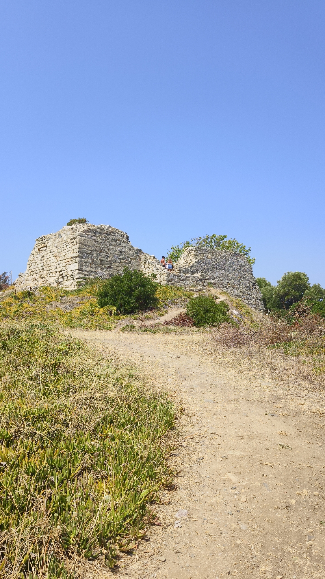 The image depicts a sunny outdoor scene featuring ancient stone ruins located in the Municipal Unit of Toroni, Greece. A dirt path curves through the foreground, bordered by dry grass and low-lying succulent plants on the left, and dry brush on the right. In the middle ground, a section of ancient stone ruins is visible, with one prominent structure appearing to be a partially collapsed tower or wall made of large, light-colored stones. Two individuals are seated on the stone steps leading up to the ruins. Green bushes and trees are scattered around the ruins and in the background, with a clear, bright blue sky overhead. The overall impression is one of historical site exploration under good weather conditions.