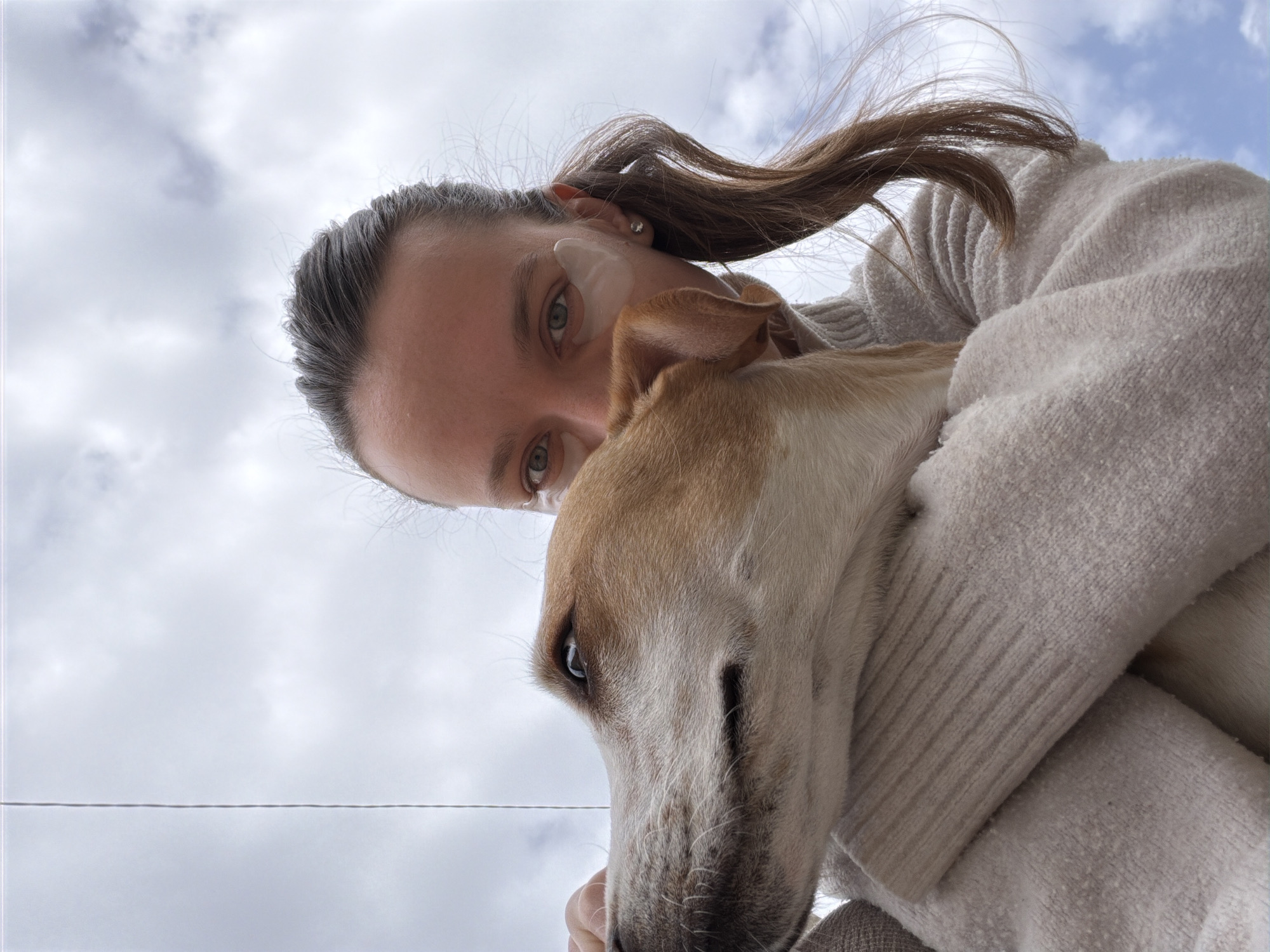 A low-angle shot depicts a person holding a light brown and white dog, likely a greyhound, against a cloudy sky. The person, fair-skinned with brown hair pulled back, is wearing a cream-colored sweater. They have what appears to be an eye mask under one eye. The dog is nuzzling into the person's sweater. A thin wire bisects the lower part of the sky. The location cannot be determined to be Budva, Montenegro, or any other specific location, based on the given image.