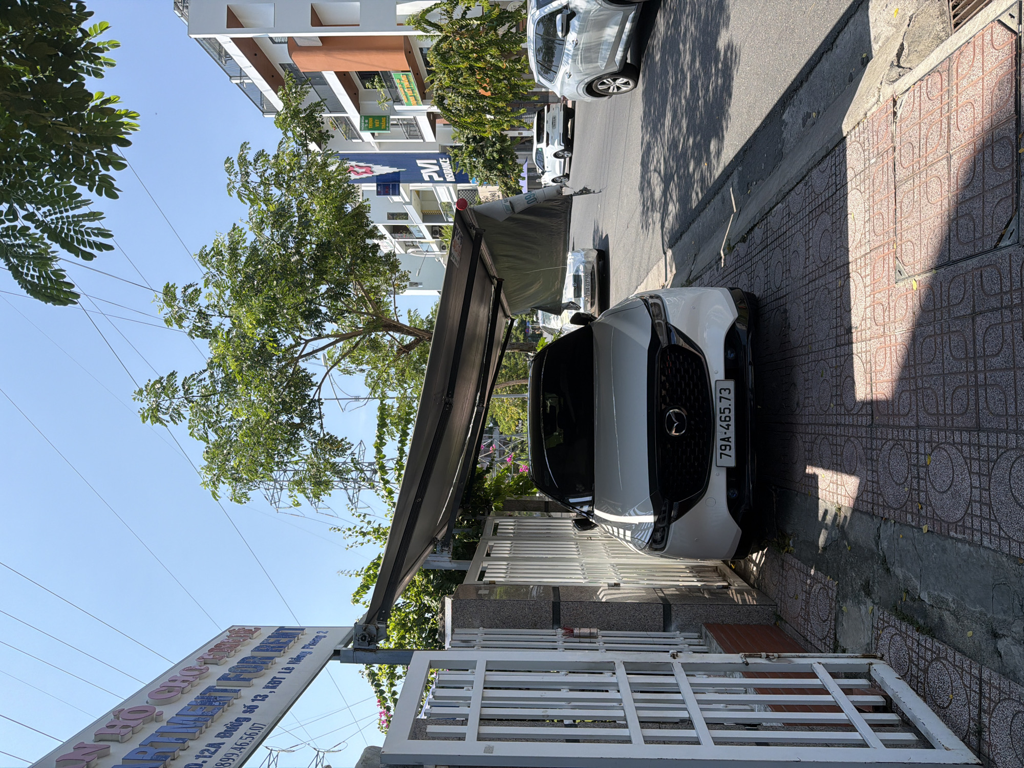 A white Mazda CX-5 is parked on a tiled sidewalk in front of a white metal gate and a granite-clad pillar. The car is partially covered by a gray awning that appears to be torn in one section. A license plate reading "79A-465.73" is visible on the front of the car. The sidewalk is paved with patterned red tiles and has a drainage grate. A street runs alongside the sidewalk, with other cars parked and driving on it. In the background, there are multi-story buildings, trees with green foliage, and a blue sky. A sign with Vietnamese text and "PVI INSURANCE" is visible on one of the buildings. The location is identified as Nam Nha Trang Ward, Vietnam.