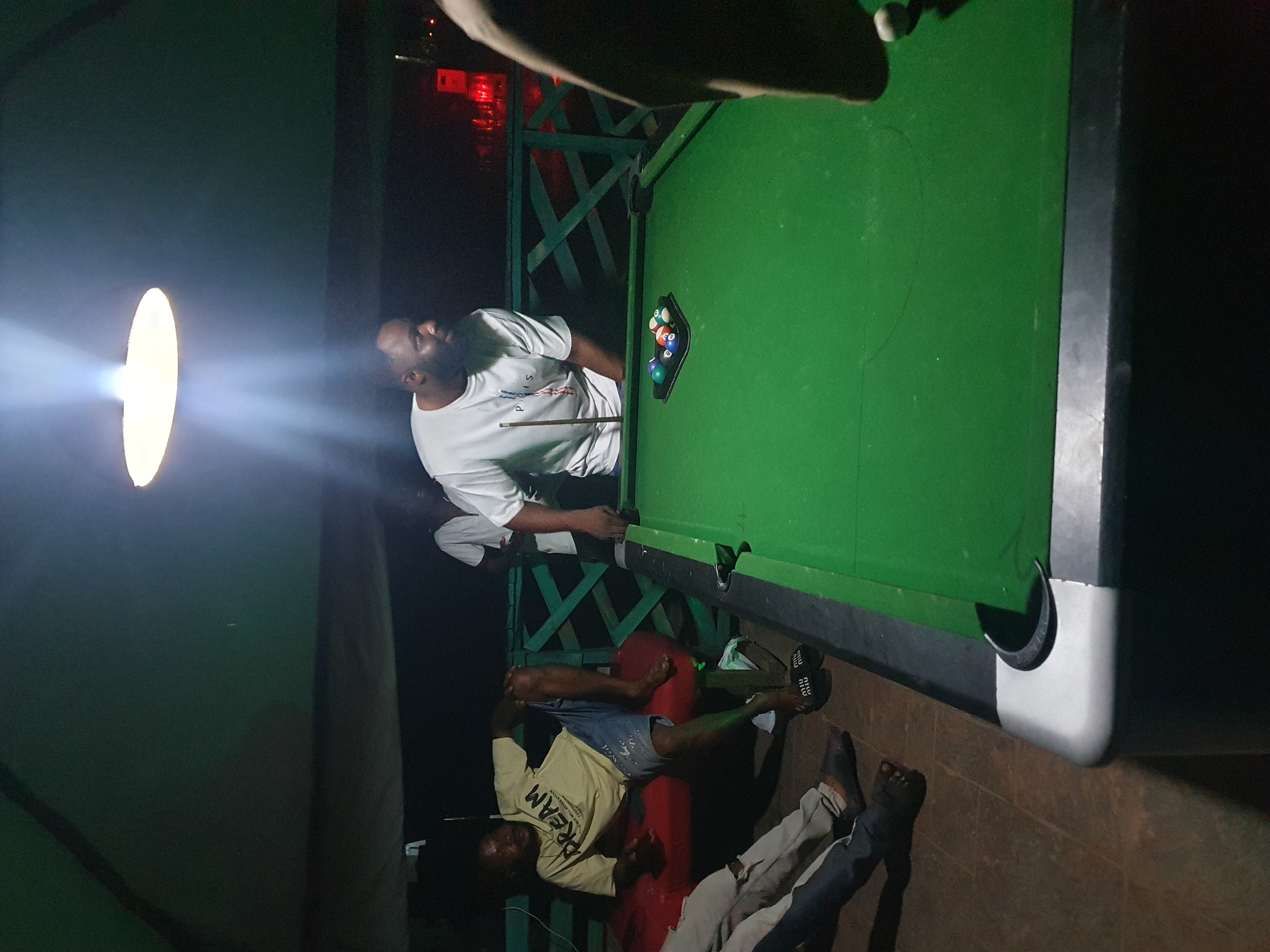 In Lokoja, Nigeria, a dimly lit outdoor setting features a pool table with several billiard balls arranged in a triangle rack. A man in a white t-shirt stands near the table, holding a pool cue. To the left, another man is seated on a red bench, wearing a yellow t-shirt and blue shorts. Two other individuals are partially visible, with one sitting and another standing, their legs and feet showing. The scene is illuminated by a bright light fixture hanging overhead, casting a spotlight on the table and the man with the cue. A green railing and a dark background with some distant red lights suggest a nighttime ambiance, possibly at a bar or recreation area.