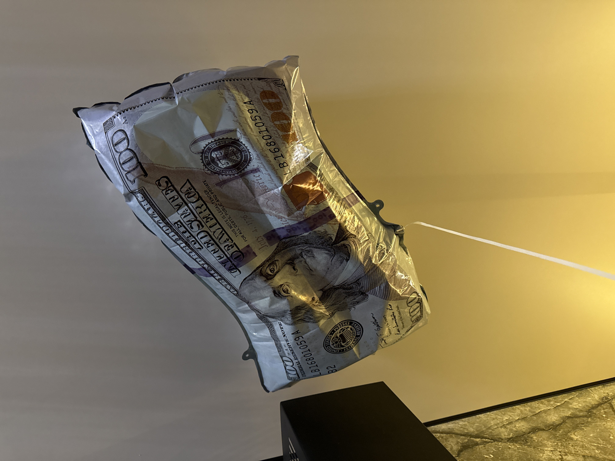 The image displays a large, metallic foil balloon designed to resemble a U.S. one-hundred-dollar bill, floating or suspended against a wall. The balloon is highly reflective, showing various creases and glares from an unseen light source. Prominent features of the $100 bill are visible, including the portrait of Benjamin Franklin, the numeral "100" in the corners, the text "UNITED STATES OF AMERICA," "FEDERAL RESERVE NOTE," and the serial number "LB16801059A." A thin, white string or ribbon extends downwards from the bottom edge of the balloon, exiting the frame. In the lower left quadrant, a portion of a dark, angular object, possibly part of a lamp or piece of furniture, is visible, next to a textured, possibly marbled, surface. The background wall transitions from a muted light gray at the top to a warm, golden yellow at the bottom, suggesting upward-directed artificial lighting illuminates the lower section. The scene is located in the city of Berezyna, Ukraine.