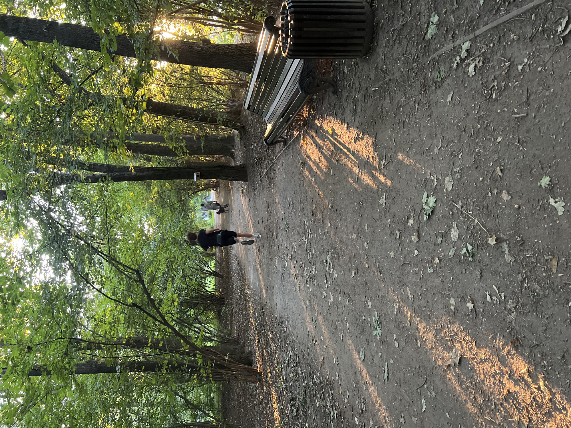 In Moscow, Russia, a park scene captures the late afternoon sun filtering through the trees, casting long shadows on a dirt path. A lone runner, wearing a dark t-shirt and shorts, moves away from the viewer down the path. Further ahead, another individual, possibly walking a dog, is visible. To the right, a park bench and a trash receptacle stand beside the path. The surrounding environment is characterized by dense foliage and mature trees.