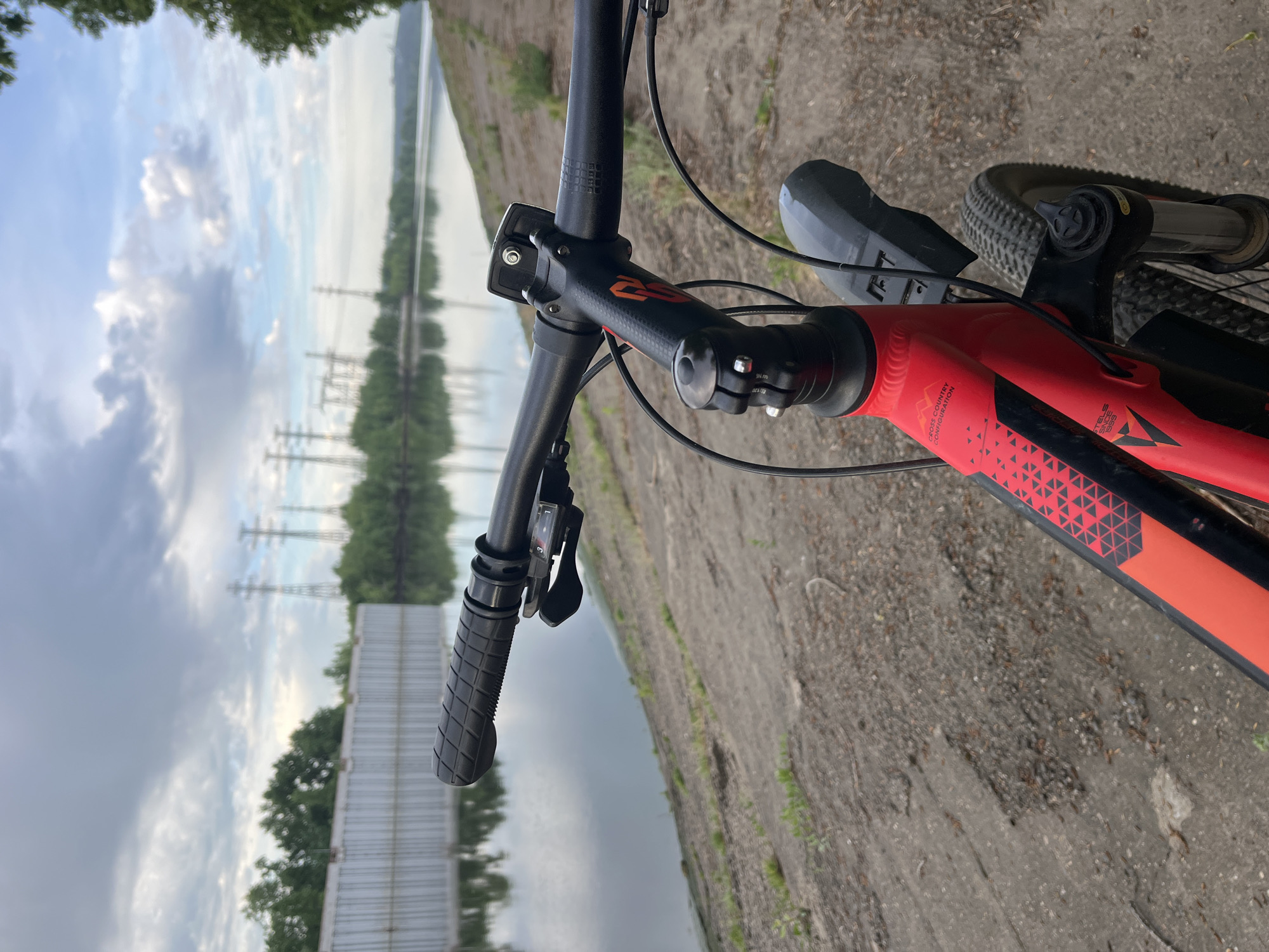 The image shows a close-up of a bicycle's handlebars and front frame, positioned in an outdoor environment. The bike is red and black with a triangle pattern on the frame. The handlebars are black with black grips and gear shifters. The front wheel and a partial mudguard are visible. The background reveals a body of water, possibly a river or a lake, with overcast skies reflected in the water. Power lines are also visible in the distance, suggesting an industrial or semi-urban setting. The foreground consists of an unpaved surface, likely a dirt road or path. Without specific landmarks or information, definitively pinpointing the exact location in Chaikovsky, Russia is impossible, but the presence of power lines and a large body of water suggests proximity to industrial or infrastructural elements near the river.