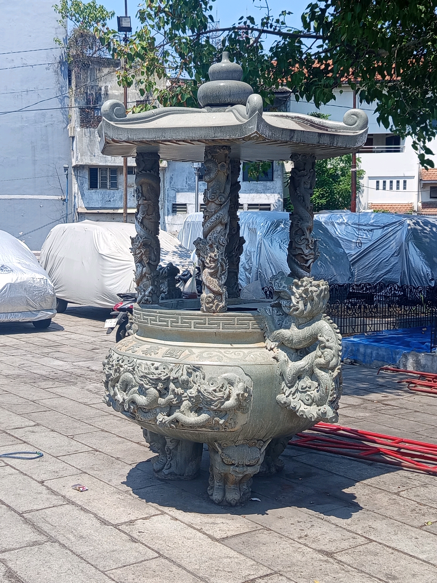 In Semarang, Indonesia, a large, ornate stone censer stands prominently in an outdoor plaza. The censer is intricately carved with dragon motifs and rests on four lion-like feet. It is a substantial piece, possibly made of granite, with multiple tiers and a decorative roof.
The plaza is paved with square stone tiles. In the background, several vehicles are covered with white tarps, suggesting they are either parked for storage or undergoing maintenance. A motorcycle is also partially visible between two of the tarp-covered vehicles. Further back, buildings with windows are visible, some with a light blue facade and others with white walls and red tiled roofs. Trees with lush green foliage are scattered throughout the scene, casting shadows on the ground.
The environment appears to be an open, urban space, possibly a courtyard or a public area adjacent to buildings. The sky is clear and blue, indicating a sunny day. There are no people directly interacting with the censer or the vehicles. However, a few objects are scattered on the ground, including a blue rope and a small pink and white item, possibly a cigarette pack. Red metal bars, possibly part of temporary fencing or scaffolding, are also visible in the lower right portion of the image. The overall impression is one of a quiet, possibly historical or cultural site within a city.