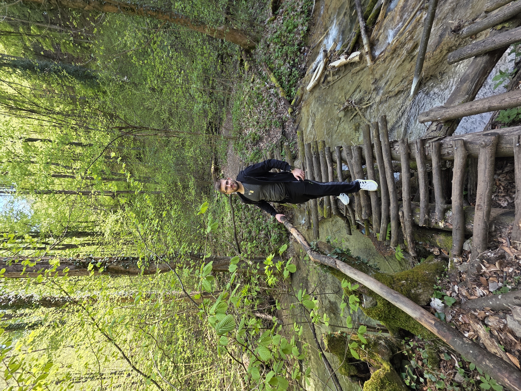 A man is standing on a wooden bridge in a forest. The bridge spans a stream. He is wearing a black jacket, a gray shirt, and black pants, and white sneakers. He is looking at the camera. The forest is green with lush trees. The water appears clear. The location is near Бачковски манастир, Bulgaria.