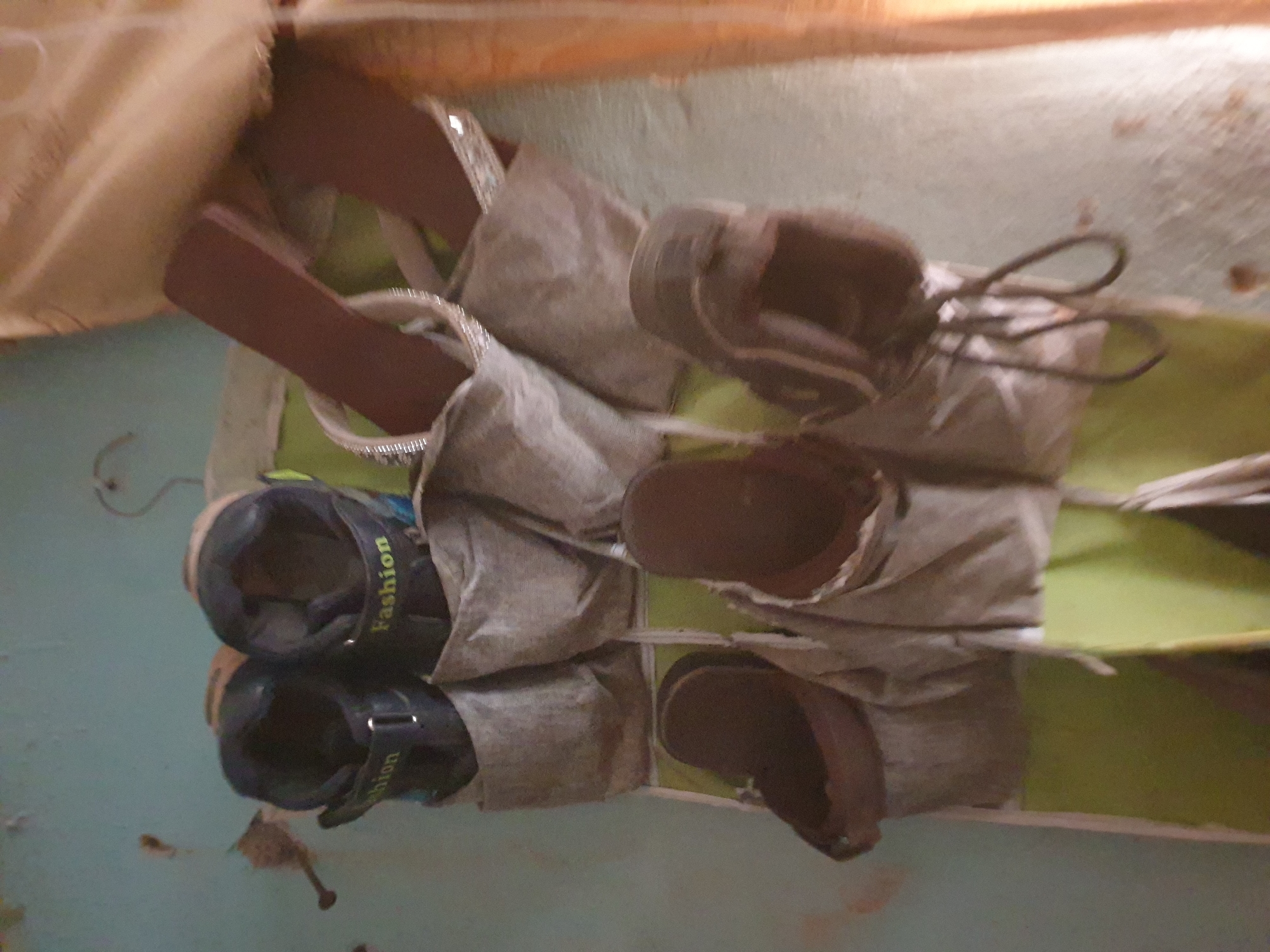 The media file shows a shoe organizer with several pairs of shoes. The organizer is made of fabric and is hanging on a wall. There are at least three pairs of shoes visible. One pair of black sneakers has the word "Fashion" printed in yellow on the side. Another pair appears to be brown boots. The scene is dimly lit, suggesting it might be indoors or nighttime. The background is a light blue wall. The shoes are in various states of wear, indicating they are used. The overall impression is of a functional but somewhat cluttered storage solution. The location context is Jalingo, Nigeria.