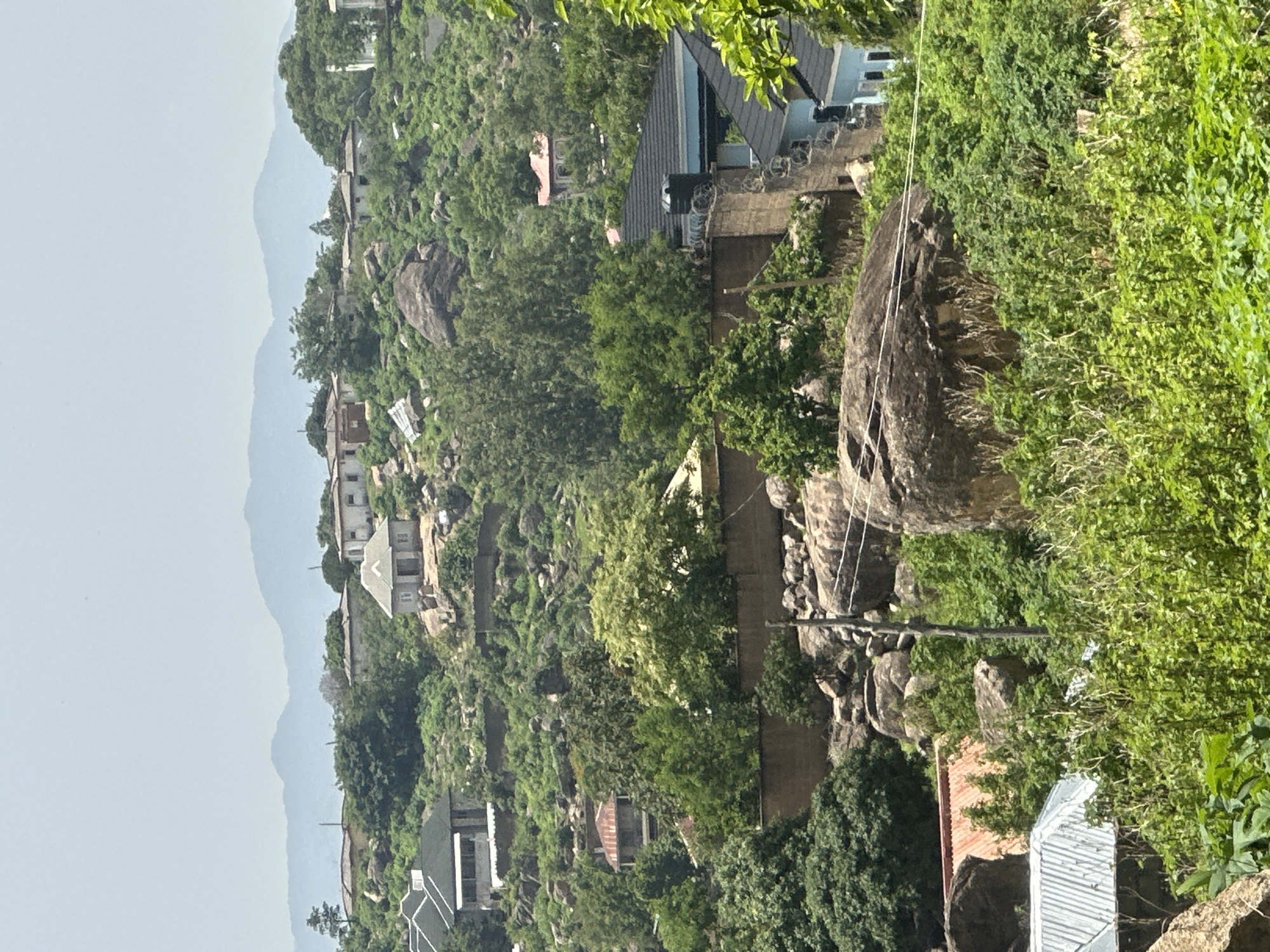 The image depicts a residential area nestled on a hilly terrain in Jos, Nigeria. The landscape is characterized by a mix of rocky outcrops and lush green vegetation, with houses strategically built into the slopes. Several structures are visible, ranging from smaller, single-story dwellings with light-colored walls and dark roofs, to larger, multi-story homes featuring a variety of architectural styles. Some houses have white or light-colored facades, while others are painted in darker hues. The roofs are predominantly dark, with some appearing to be made of metal or tile. Walls of rough-hewn stones and concrete are used to terrace the hillsides and delineate properties. The vegetation is dense, with numerous trees and shrubs contributing to the verdant appearance of the scene. In the background, distant mountains are visible under a hazy sky, suggesting a broad panorama. No people are present in the frame. The overall impression is of a community integrated with its natural, rocky environment.