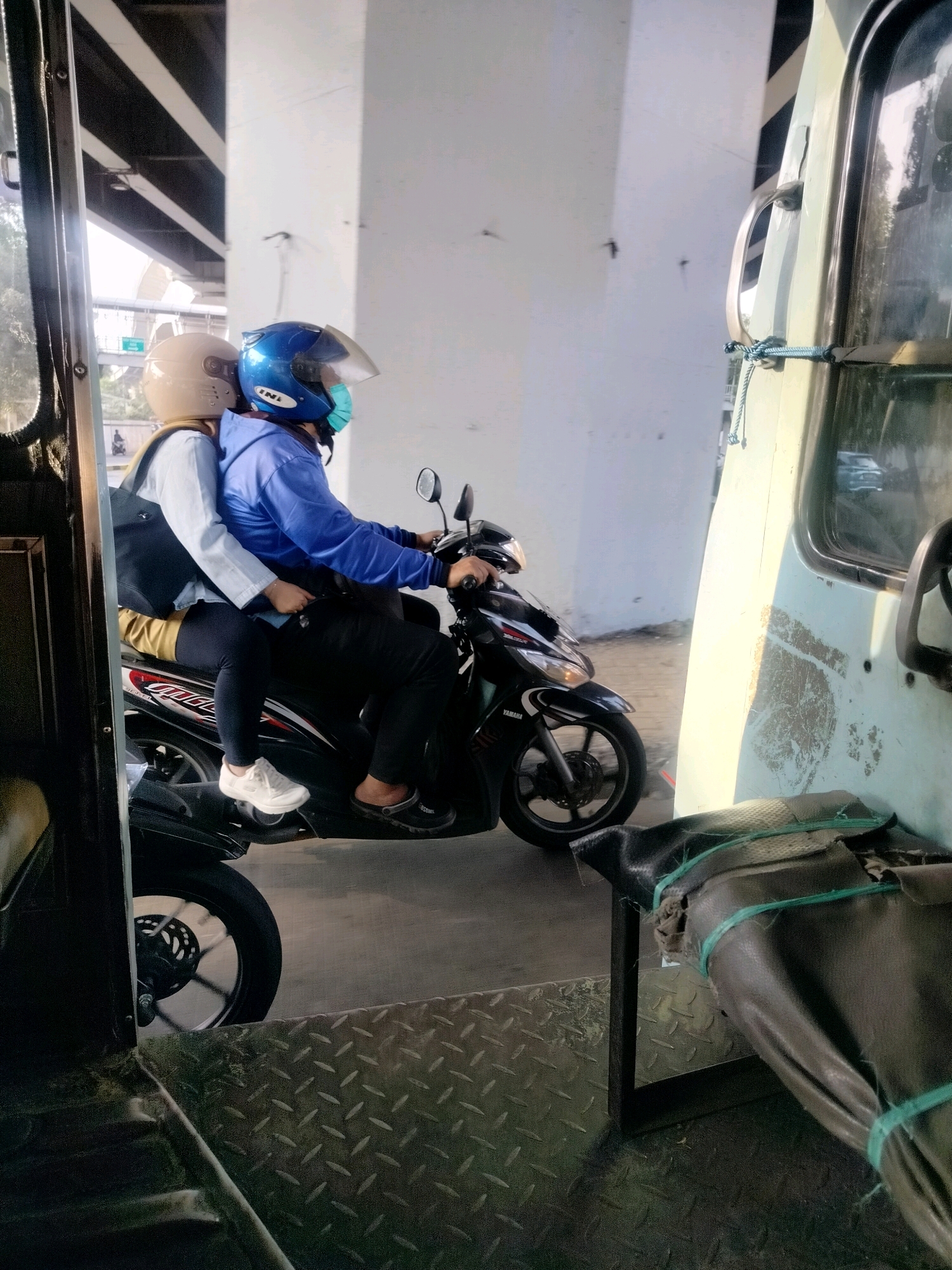 Two individuals, wearing helmets and face masks, are riding a motorcycle. The person in the front, likely the driver, is wearing a blue long-sleeved shirt and black pants. The person in the back, the passenger, is wearing a light-colored long-sleeved shirt, yellow pants, and a black backpack. They appear to be in traffic, possibly on a flyover or elevated road, with a concrete structure and overhead beams visible in the background. A portion of a light-colored vehicle, possibly a bus, is to the right of the motorcycle, with a metal handle and a rope securing an object to its side. The ground is a textured metal surface. This scene is consistent with traffic conditions in the city of East Jakarta, Indonesia.