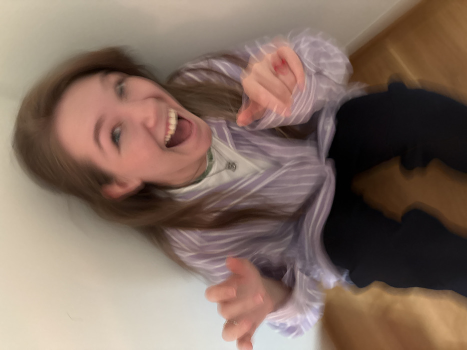 A single individual, appearing female with long brown hair, is depicted in an indoor corner. She is positioned on a light-colored wooden floor against plain white walls. The individual wears a white undershirt, a light purple and white striped long-sleeved button-up shirt, and dark trousers. A necklace with light green beads and a silver-colored pendant is visible. A ring is present on the ring finger of the left hand, and red nail polish is visible on the right hand. The individual is captured mid-action, with a wide-open mouth suggesting laughter or a shout, and eyes wide open. Her left hand is raised with fingers splayed, and her right hand is extended forward, pointing or gesturing. Significant motion blur is evident across the entire subject, particularly on the face and hands, indicating movement during image capture. The scene is an interior space with no discernible external geographic identifiers. The user specified the city of Belgrade, Serbia.