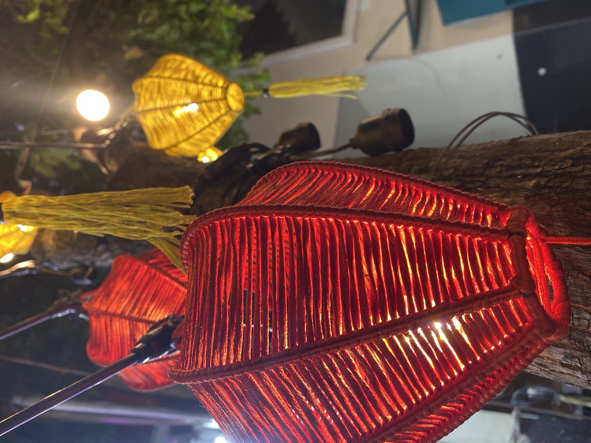 This is a close-up, low-angle shot of several lanterns hanging from a tree at night, likely in the Son Tra district of Đà Nẵng, Vietnam. The lanterns are woven from red and yellow twine, with the red lantern in the foreground prominently displaying the warm glow of an internal light source. Yellow tassels hang from the yellow lantern in the background. The tree trunk is visible, providing a textured backdrop, and a bright white light bulb is also visible, suggesting outdoor lighting. The overall ambiance is festive and slightly mysterious due to the low light conditions and focus on the illuminated lanterns.