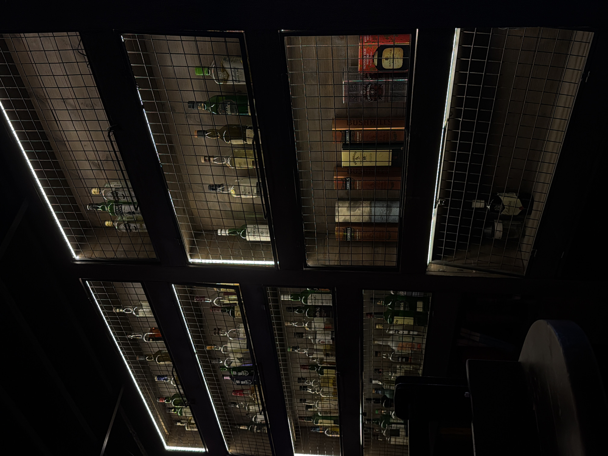This image shows a ceiling display of liquor bottles and boxes behind a metal grid, illuminated by strips of light along the frame. The bottles and boxes appear to be arranged within individual compartments or sections. Brands visible include Glenfiddich, Glenlivet, Laphroaig, Bushmills, Chivas Regal, and Suntory Old. Part of a dark gray round structure is visible in the foreground, obscuring the lower right corner of the display. The overall lighting is subdued, with the light strips accentuating the display's structure. It is impossible to verify if this is Izhevsk, Russia without additional information.