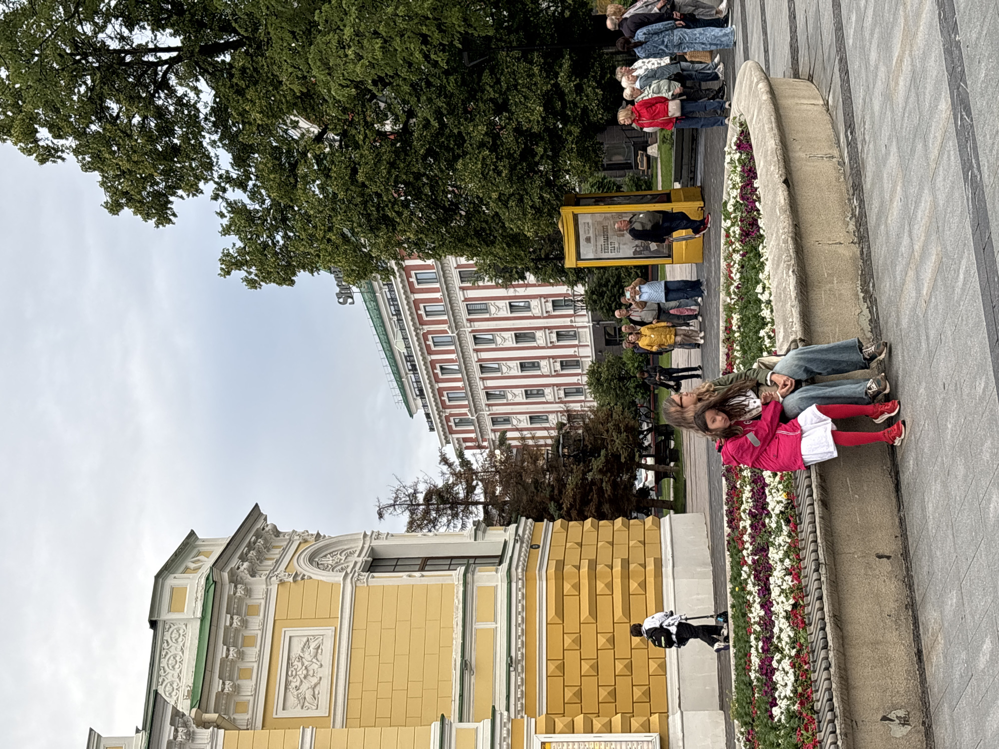 This image captures a street scene in Nizhny Novgorod, Russia, featuring a building with yellow facades and intricate white architectural details in the foreground, possibly a historical landmark or theater. To the right of this building, a modern hotel with red and white striped facades and the "Sheraton" logo is visible, nestled amidst lush green trees. In the mid-ground, a diverse group of people are gathered, some looking towards the hotel, while others are engaged in conversation or taking photographs. A prominent yellow advertising kiosk stands near the hotel entrance. In the foreground, two individuals, a woman and a young girl, are seated on a bench by a flowerbed filled with white and purple flowers. The girl is wearing a bright pink jacket and white dress, while the woman is dressed in a green jacket and jeans. The pavement is paved with large gray tiles. The overall atmosphere appears to be that of a public square or street in a city during the daytime, with overcast skies suggesting a mild weather day. A person on a scooter is seen in the background, adding to the dynamic nature of the urban environment.