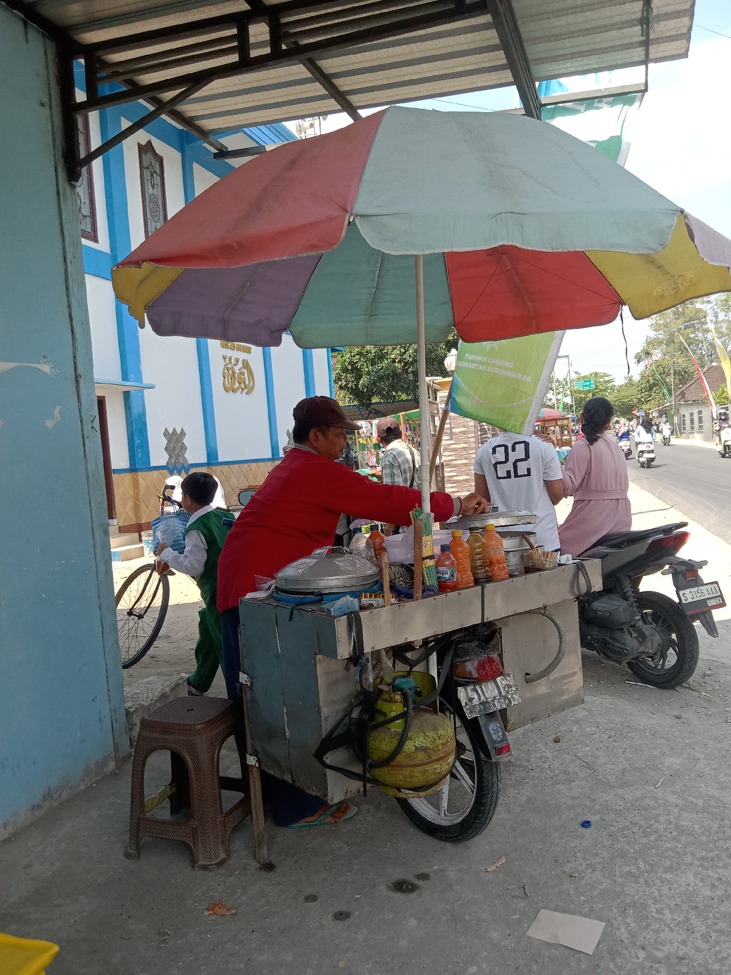 In Sugihwaras, Indonesia, on a sunny day, a street vendor is serving food from a cart parked next to a road. The cart is colorful, adorned with a large, multi-colored umbrella, and mounted on a scooter. Several bottles of beverages are displayed on the cart's counter. A woman in a pink dress and a man in a white t-shirt with the number "22" on the back are customers at the cart. A young boy in a green and white uniform is standing with a bicycle. In the background, a mosque with blue and white architecture is visible, along with other people and vehicles on the street.