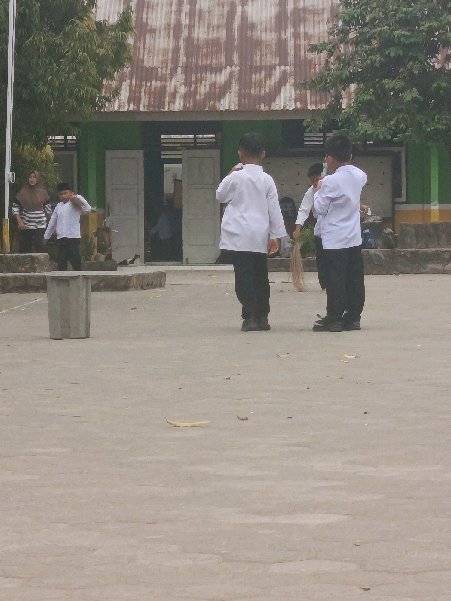 The image depicts an outdoor scene in front of a low-rise building with a corrugated metal roof, possibly a school. Several children, appearing to be of school age, are visible. Two boys in the foreground are wearing white long-sleeved shirts and dark pants. One of them is holding a broom made of dried palm fronds. Another child is seen in the background, also in a white shirt and dark pants, holding a broom. A woman wearing a hijab and a striped shirt stands to the left, observing the children. The ground is paved with concrete. The environment appears to be a yard or open space adjacent to the building. The overall lighting suggests it is daytime, possibly overcast.