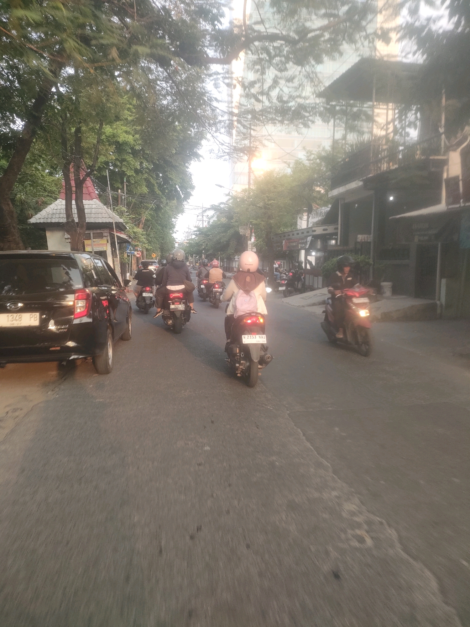 In Surabaya, Indonesia, a street scene unfolds at dusk, characterized by a line of motorcycles and a car navigating a paved road bordered by buildings and trees. A black SUV, license plate "L 1348 PB," is prominent on the left. Several motorcycles follow, their riders adorned with helmets and some with backpacks. One rider in the foreground wears a light pink hijab and backpack, on a motorcycle with license plate "L 2153 NAI." The scene suggests ordinary daily life and traffic on a city street.