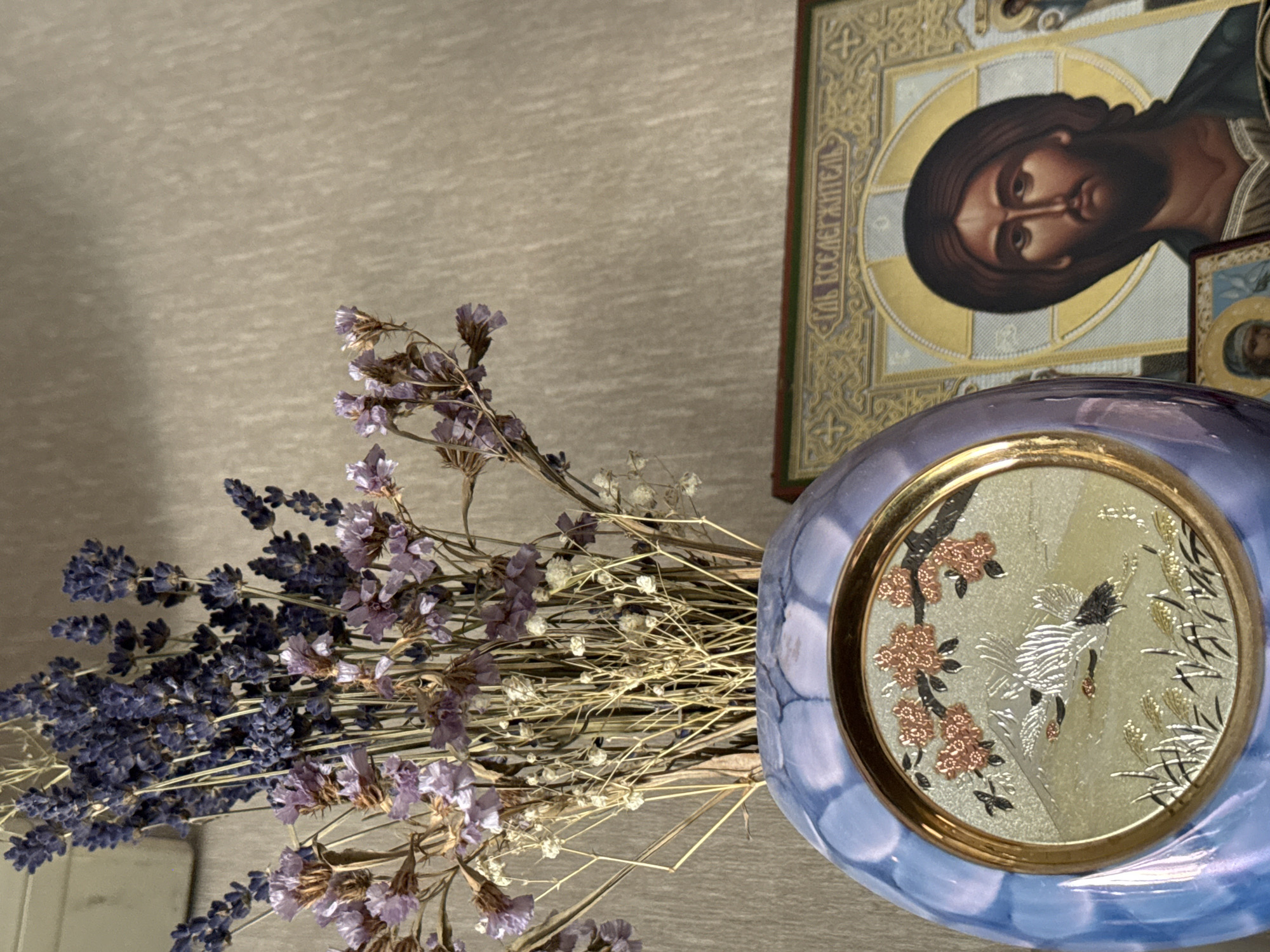 A close-up shot shows a vase with dried lavender and other flowers. To the right of the vase is an icon of Jesus Christ, with smaller icons placed below it. The background is a textured beige wall. The scene appears to be indoors, possibly in a home or religious setting in Bishkek City, Kyrgyzstan. There are no people present in the image.