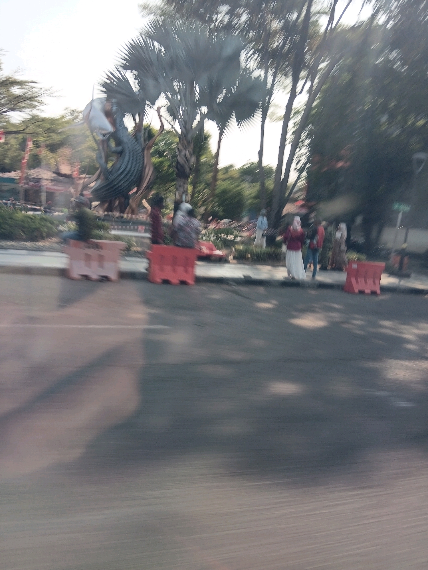 This is a street-level, medium-range shot of a public area, likely a park or a plaza, in Surabaya, Indonesia. The image is characterized by motion blur, suggesting it was taken from a moving vehicle.
In the background, a large sculpture resembling a dragon or mythical creature dominates the scene. The sculpture is dark grey with metallic accents and appears to be intertwined with tree trunks and branches. Behind the sculpture, lush green foliage and trees are visible, with a few buildings peeking through the dense greenery.
In the mid-ground, several individuals are present. Some are standing near the sculpture, while others are seated on or near orange and white concrete barriers that line the edge of the road. The people are dressed in casual attire, with some individuals wearing headscarves. The barriers are positioned to create a physical separation between the road and the pedestrian area.
The foreground is largely occupied by a blurred asphalt road surface, indicating the perspective of a vehicle in motion. Shadows from trees and possibly the sculpture are cast on the road, creating a dappled effect. The overall lighting suggests it is daytime, with bright sky and sunlight filtering through the trees. The presence of barriers and the sculpture suggests this may be a public space designed for leisure and cultural appreciation.