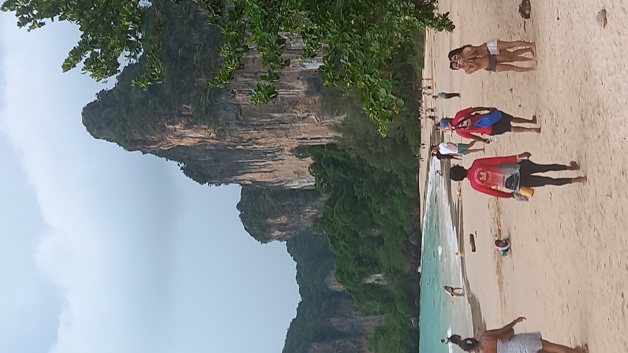 The image captures a beach scene in Ban Ao Nang, Thailand, set against a backdrop of towering cliffs covered in lush vegetation. The sky is overcast with grey clouds. A narrow strip of turquoise water separates the sandy beach from the cliffs.
On the beach, a number of people are visible. Two individuals are lying close together, and another group is standing nearby. Several figures wearing red shirts, possibly lifeguards, are positioned along the shoreline. A few others are walking or standing near the water's edge. The sand is light in color and appears to be packed down.