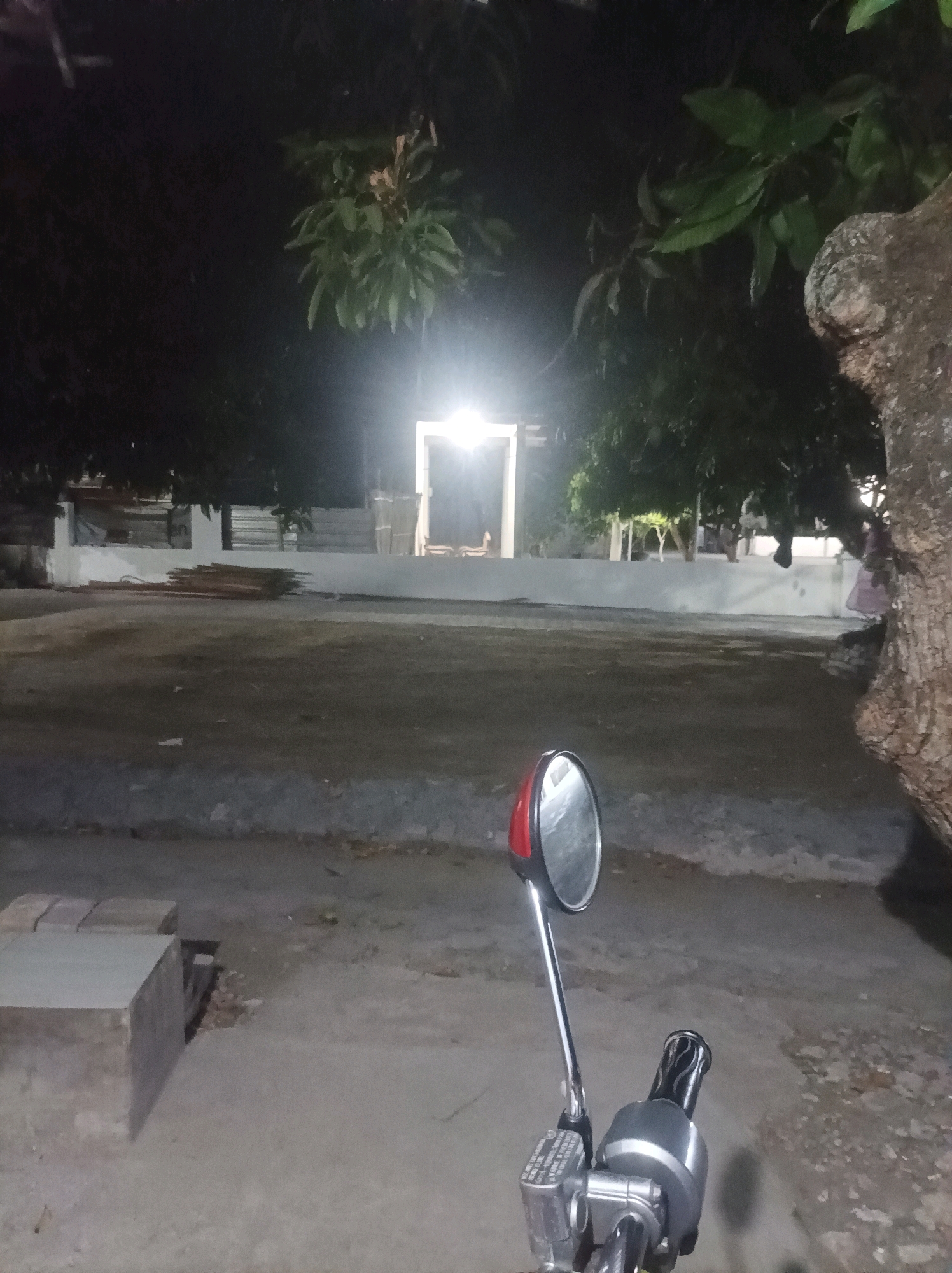 This image was taken at night in Canga'an, Indonesia. The scene is dimly lit by a bright light source in the background, which illuminates a doorway or archway. In the foreground, part of a motorcycle is visible, including a handlebar with controls and a rearview mirror. The motorcycle is parked on a paved area. Beyond the motorcycle, there is a dirt or gravel ground and a low white wall. Trees with dark foliage are visible overhead and to the right, with the textured bark of a large tree prominent on the right side of the frame. The overall impression is one of an outdoor, possibly rural or semi-rural, setting at night.