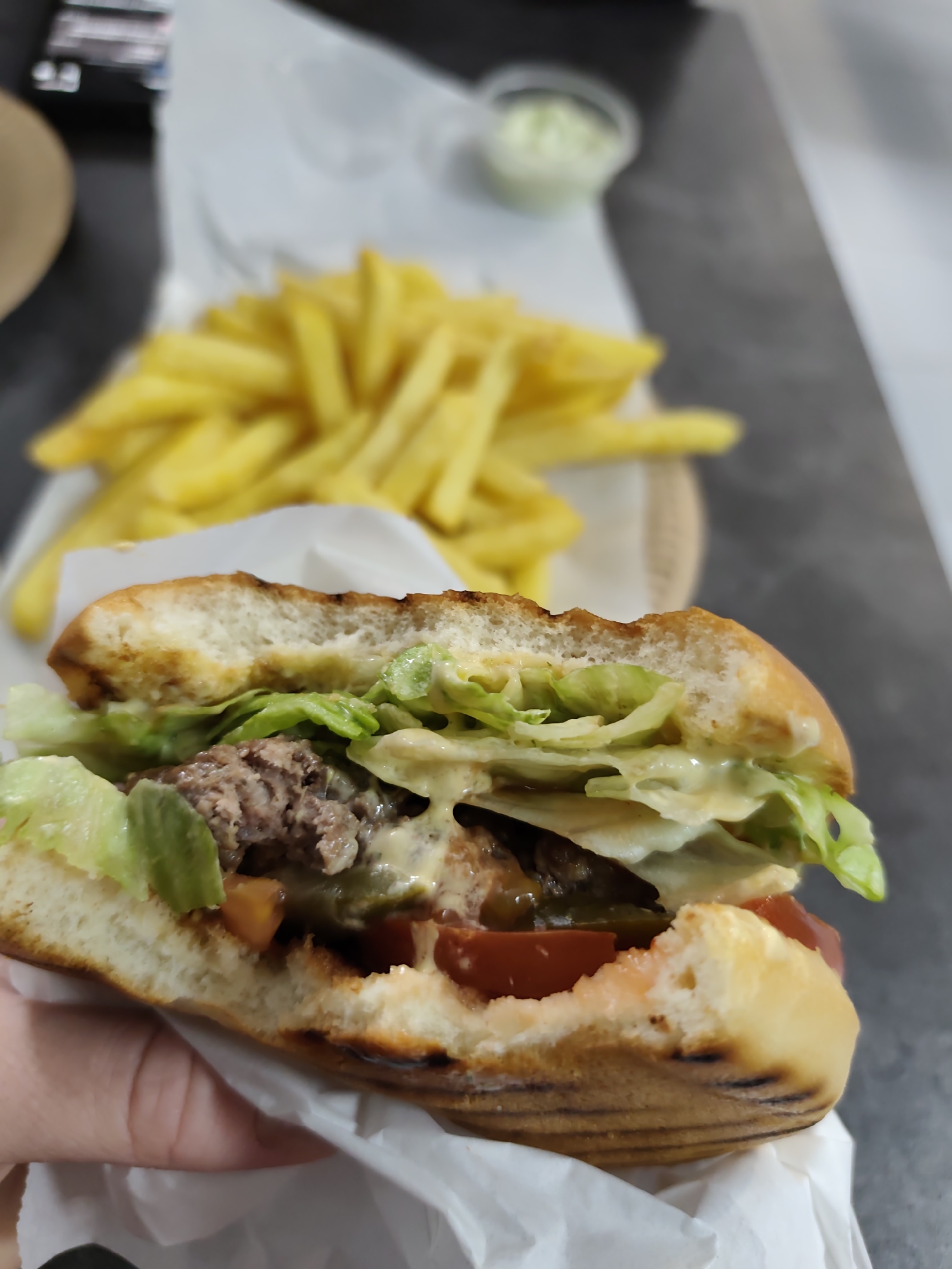 The image shows a partially eaten burger held in a hand, set against a blurred background featuring french fries and a small container of sauce. The burger includes lettuce, tomato, and a meat patty with sauce. The bun shows grill marks and a bite is missing. The setting appears to be indoors, possibly a restaurant or home. The precise location within Chaikovsky, Russia cannot be determined from the visual information provided.