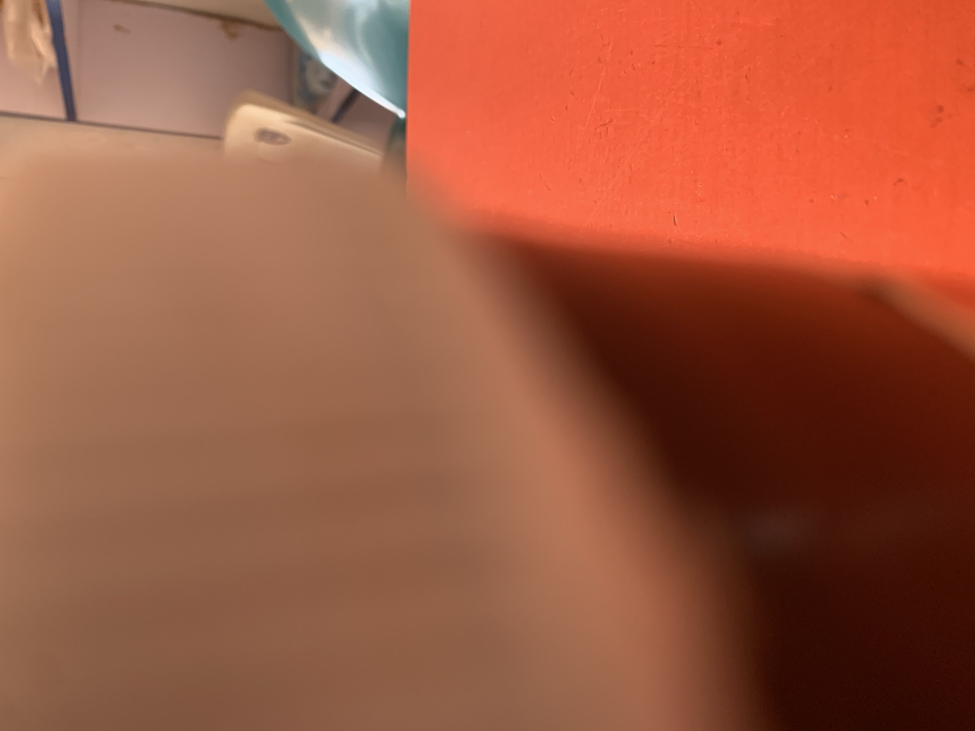 The image is a close-up, out-of-focus shot of an orange surface in the foreground. In the background, a blurry white and pink object is visible, along with a shelf with what appear to be some items on it. The overall scene is indistinct and lacking in clear detail, making it impossible to determine the specific location or context.