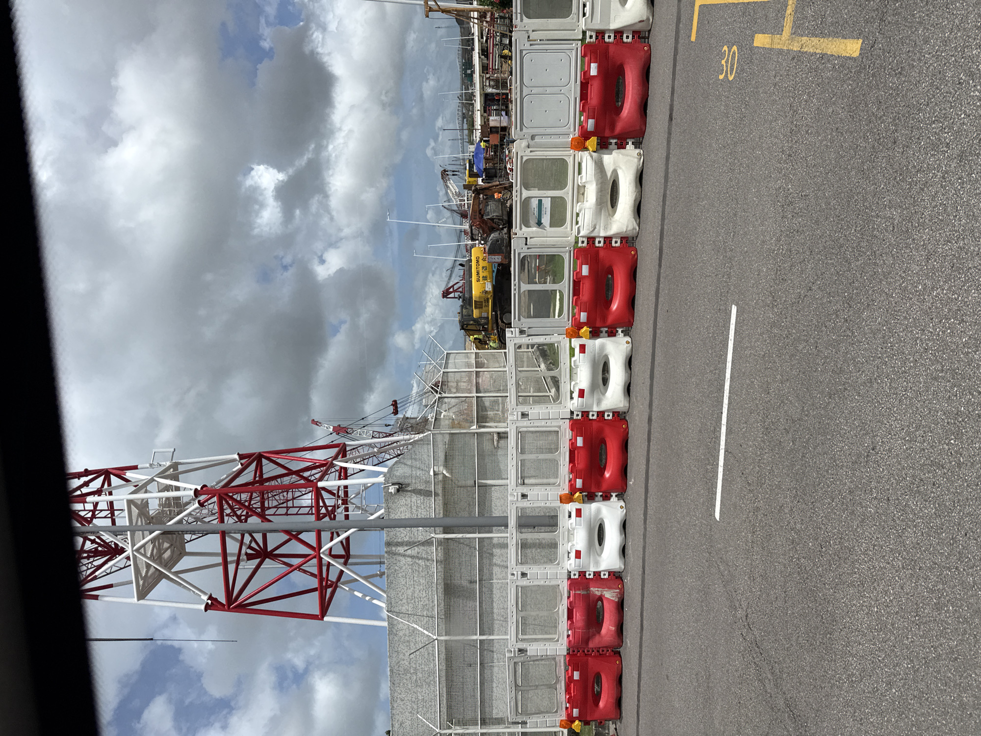 The image depicts a construction site in Macau, China, with a large red and white lattice tower dominating the background. In the mid-ground, a row of red and white plastic water-filled barriers, topped with white modular fence panels, separates the viewer from the construction area. Beyond the barriers, various construction vehicles, including a yellow Sumitomo excavator and several cranes, are visible. The environment appears to be a port or industrial area, with the water and sky suggesting an outdoor setting. The road in the foreground has painted markings, including a white line and yellow numerals indicating "30". The sky is partly cloudy with dramatic cloud formations.