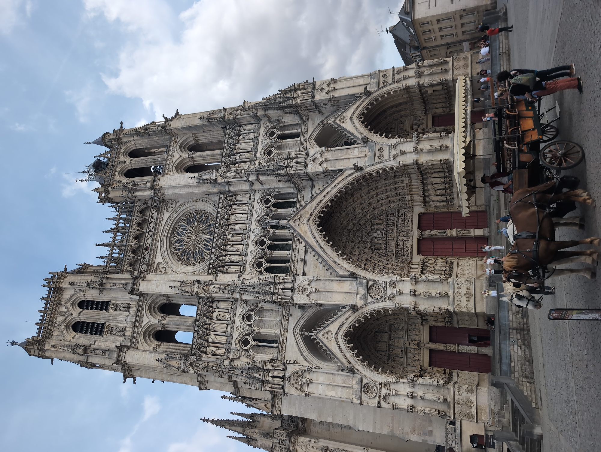 This is a low-angle, full shot of the Cathédrale Notre-Dame d'Amiens, a Gothic cathedral located in Amiens, France. The facade of the cathedral, made of light-colored stone, dominates the frame. It is highly ornate, featuring multiple tiers of sculptures, arched doorways, and a large rose window. Two prominent towers rise on either side of the central section, topped with spires. The sky above is partly cloudy. In the foreground, a horse-drawn carriage with a yellow canopy is parked on a cobblestone square. Two horses are harnessed to the carriage, and several people are visible around it, some appearing to be tourists or locals. A few other individuals are seen on the steps leading up to the cathedral and in the background, near adjacent buildings. The overall scene depicts a public space in front of a historical landmark on a cloudy day.
