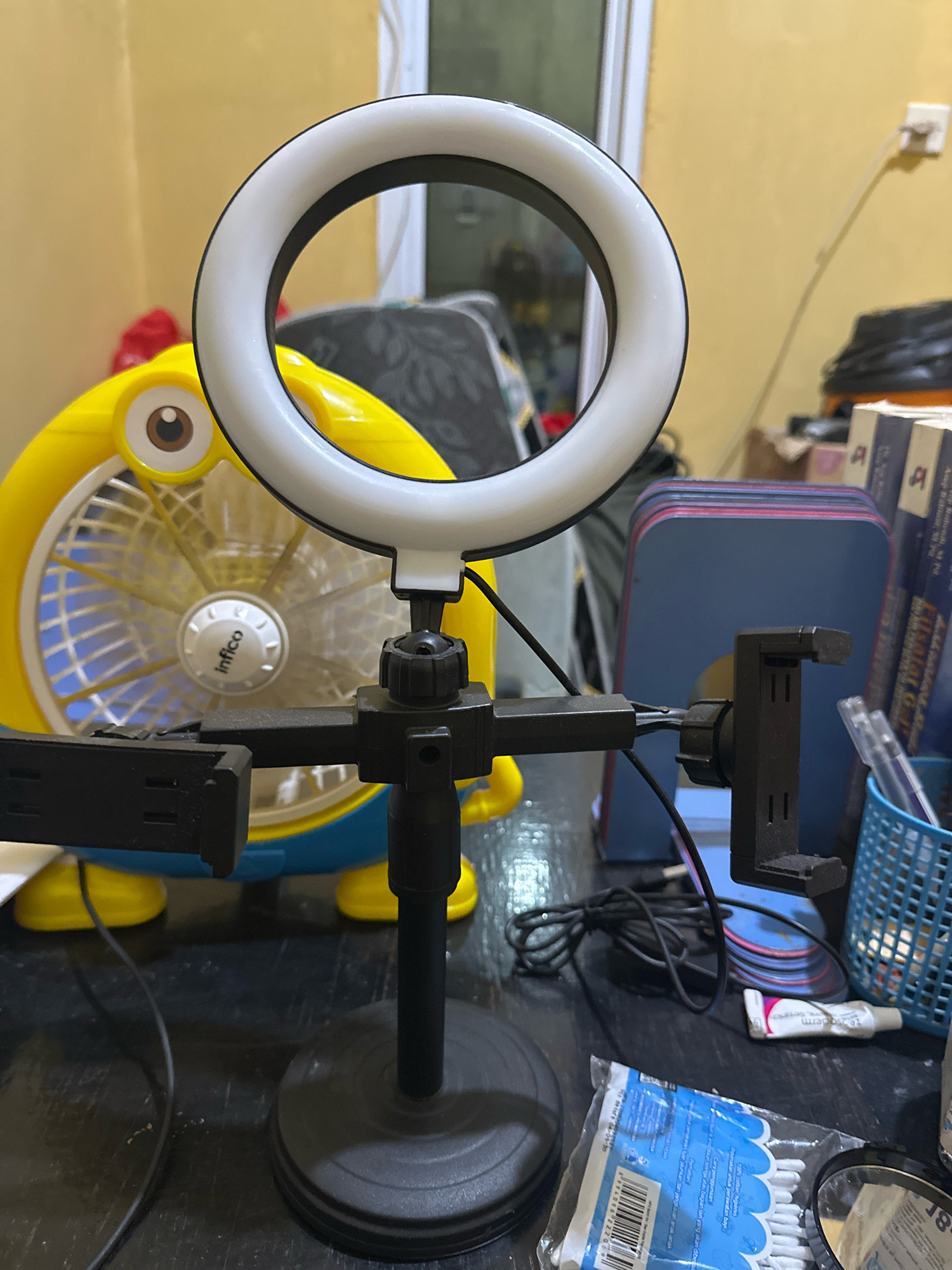 The image shows a close-up view of a selfie ring light and phone mount setup on a table. Behind the ring light, a yellow cartoon-themed fan with a large eye is visible. To the right of the ring light, there are stacks of blue and red binders, a blue basket containing pens, and a tube of cream. In the foreground, a black stand supports the ring light and phone mount. A black power cord runs from the ring light setup and rests on the table. A package of cotton swabs is also on the table in front of the phone mount. The background appears to be a room with a yellow wall and some furniture. The image is well-lit, highlighting the objects in the foreground. There are no people visible in the image. The location is not specified as Saruni, Indonesia.