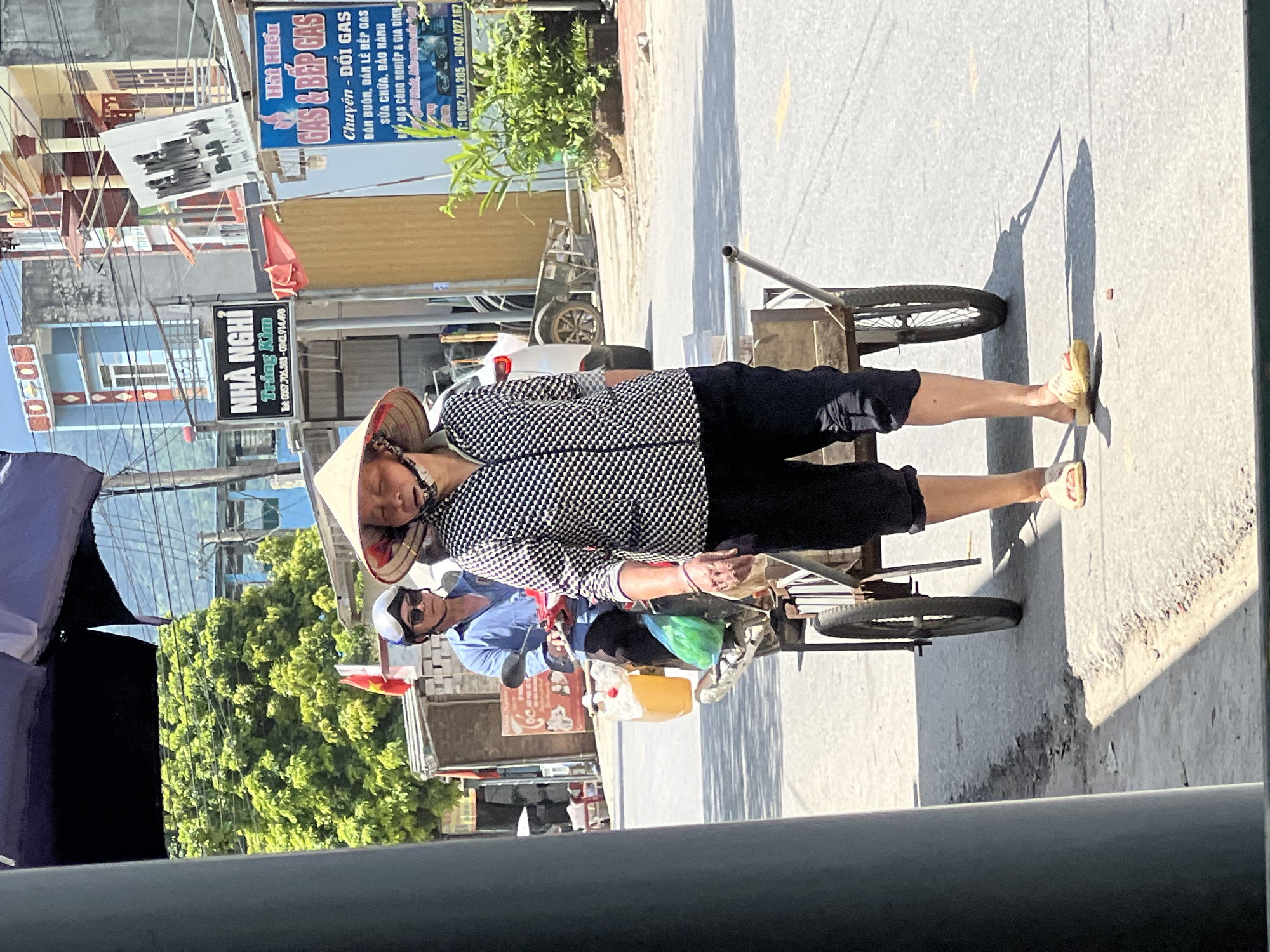 A woman wearing a conical hat and a black and white checkered shirt walks down a paved road in Lùng Tám Commune, Vietnam, pulling a wooden cart with a metal frame. She is also wearing black pants and light-colored sandals. Behind her, a man on a scooter wearing a blue shirt, helmet, and sunglasses is stopped, with a yellow plastic container and a green plastic bag on the back of his scooter. In the background, there are buildings with signs, including one for "GAS & BẾP GAS". Trees and power lines are also visible. The scene is brightly lit by sunlight, casting shadows on the road.