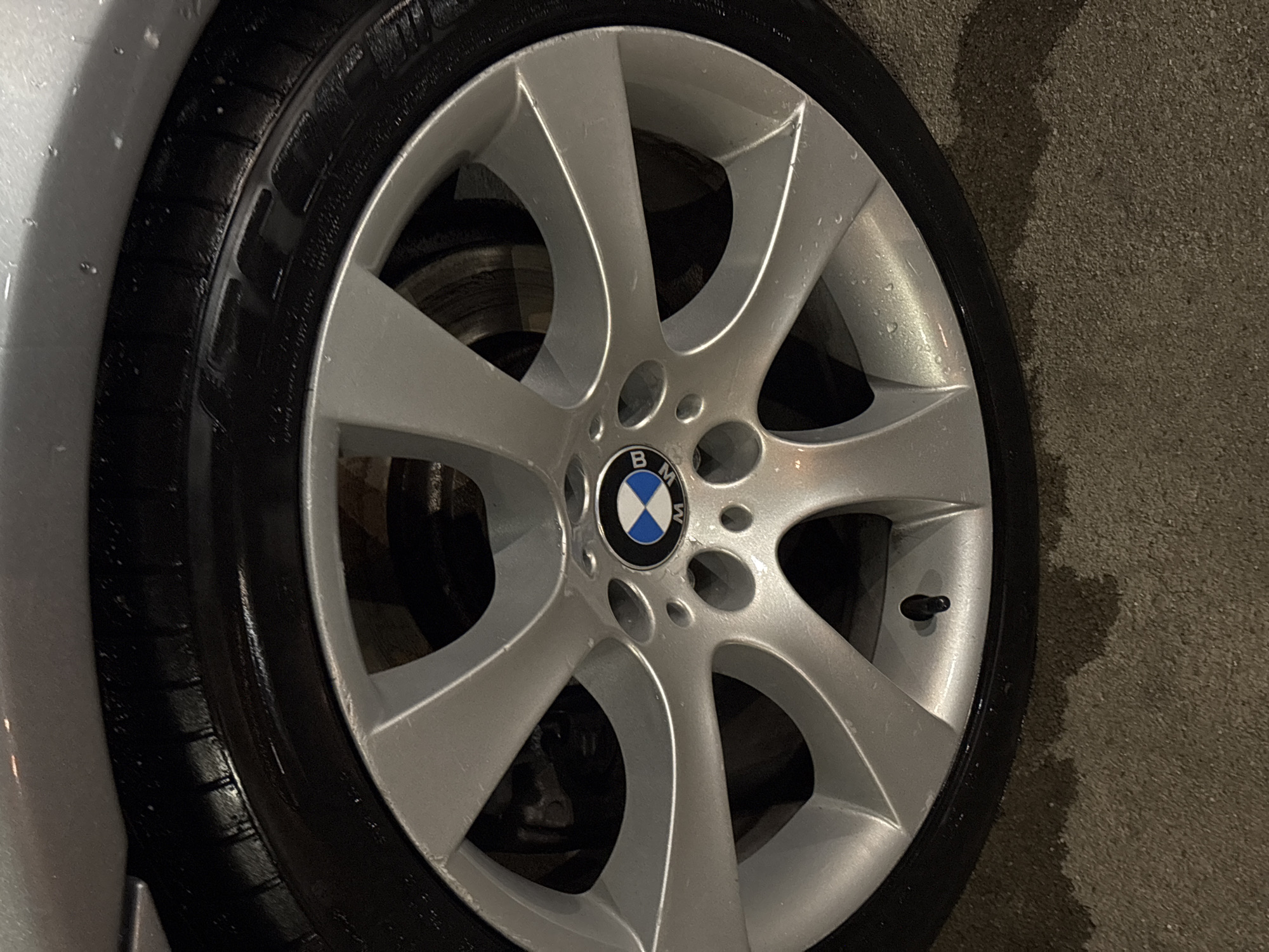 A close-up shot shows a BMW car wheel. The wheel is silver and has a BMW logo in the center. The tire is black and appears to be wet. The background is a blurred concrete surface. The location is unknown, and there is no evidence to suggest it is in Plovdiv, Bulgaria. There are no people visible in the image.