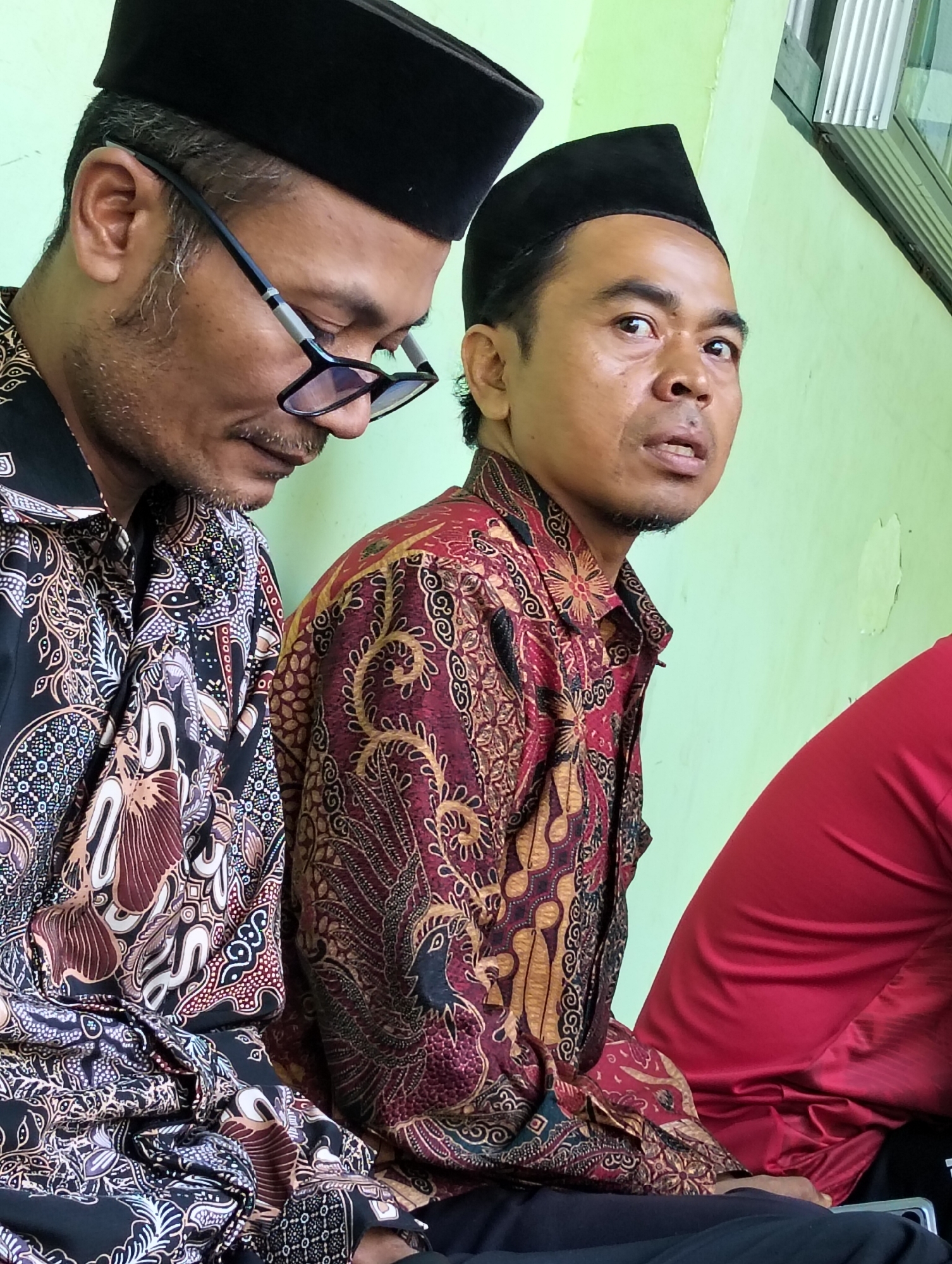 Two adult males are seated indoors, facing towards the right of the frame, against a pale green wall. Both are wearing black peci hats and batik shirts. The male on the left, partially obscured, wears glasses and a black batik shirt with brown and white patterns. The male on the right, in full view, has a patterned maroon and gold batik shirt. His expression is looking towards the camera. A third individual, wearing a red garment, is partially visible on the far right of the frame. The environment appears to be a simple room, with corrugated metal siding visible in the upper right corner. The image was taken in Dagan, Indonesia.