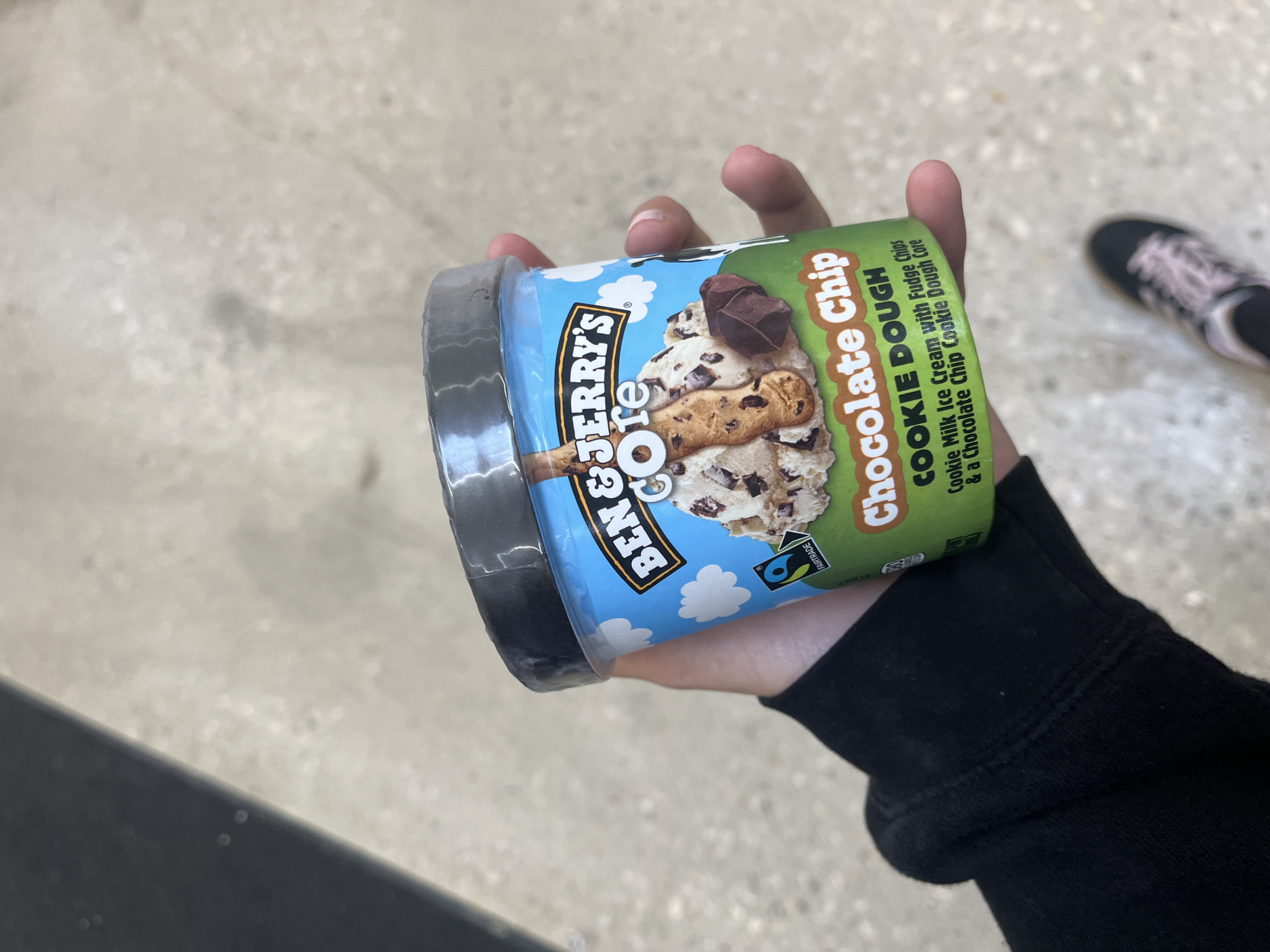 A person is holding a container of Ben & Jerry's "Core Chocolate Chip Cookie Dough" ice cream. The container features a light blue background with white clouds, the Ben & Jerry's logo, an illustration of the ice cream, and a green label with the flavor name. The person is wearing a black long-sleeved shirt. The location appears to be indoors, possibly a store, given the smooth flooring in the background. A shoe is also visible in the background. The photograph was taken in San Antonio, United States of America.