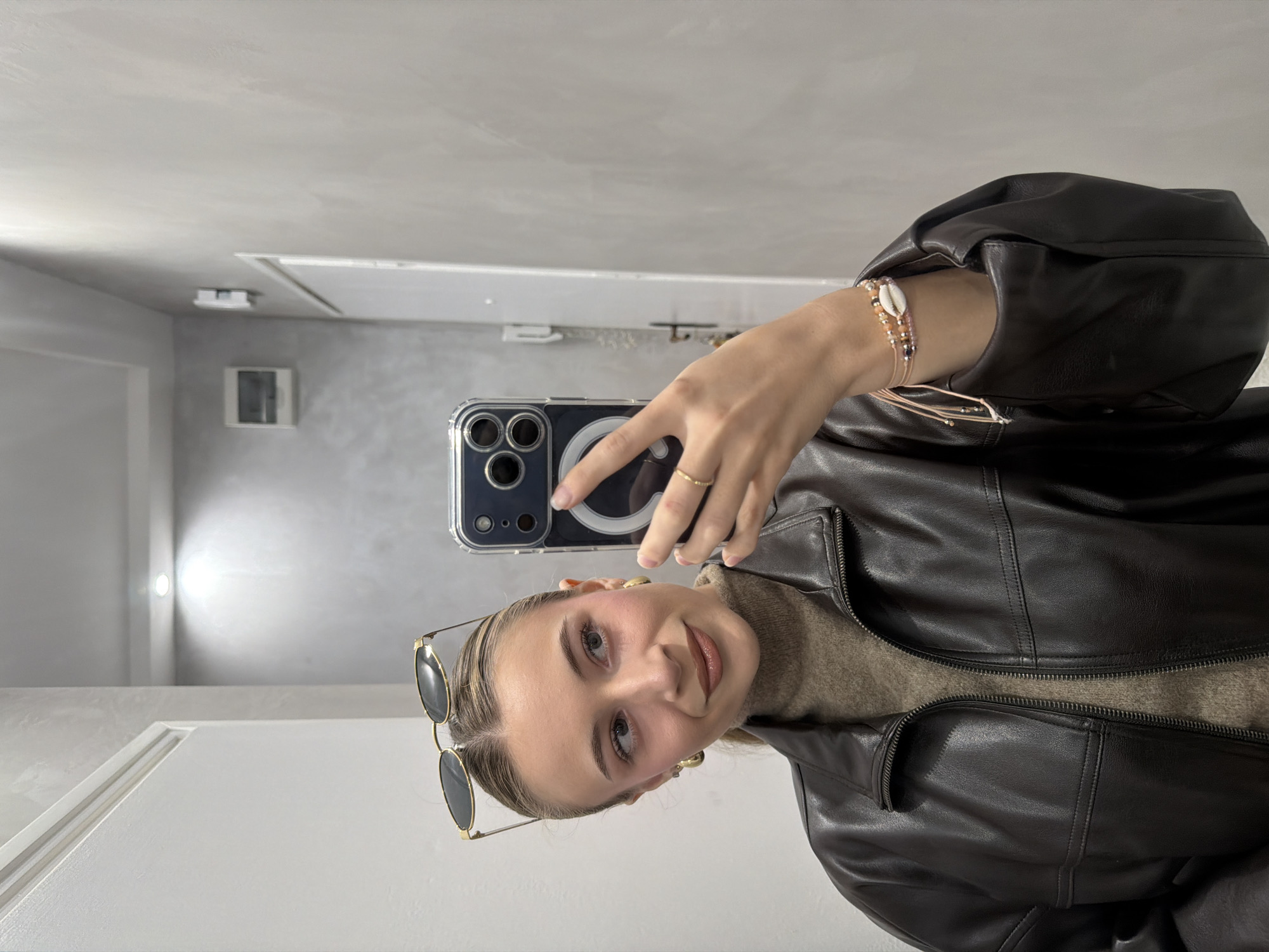 The image depicts a young adult female taking a mirror selfie inside a modern indoor space. The subject has light brown hair pulled back and wears gold-rimmed sunglasses perched on her head, along with small gold hoop earrings. She is dressed in a dark brown, possibly leather, collared jacket over a light brown turtleneck sweater. A gold ring is visible on her right ring finger, and multiple beaded bracelets, including one with a white cowrie shell and various colored beads, are on her left wrist. She holds an iPhone, visible in a clear case with a MagSafe ring and triple camera module, in her right hand, angled to capture her reflection. Her expression is a slight smile, with her gaze directed towards the phone screen. The reflected background features light grey textured walls. On the left, a white door frame is visible, and above it, a recessed ceiling light illuminates the scene. Towards the right, a partially open white door with a dark handle and a white electrical panel are visible on the wall. The ceiling is white. The precise location within Plovdiv, Bulgaria, cannot be determined from the visual details, but the setting is consistent with an urban residence or similar modern indoor environment.