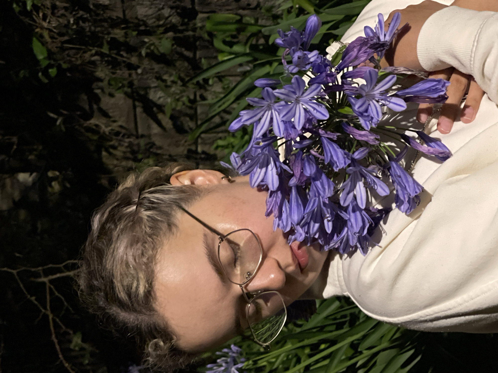 A person with short, wavy hair and glasses lies on their side, holding a bunch of purple flowers. The person is wearing a white sweatshirt. The background includes dark foliage and what appears to be a brick wall. Additional purple flowers and foliage are visible beneath the person. The location appears to be outdoors at night. The image does not provide information about the city of Lâm Viên - Đà Lạt, Vietnam.