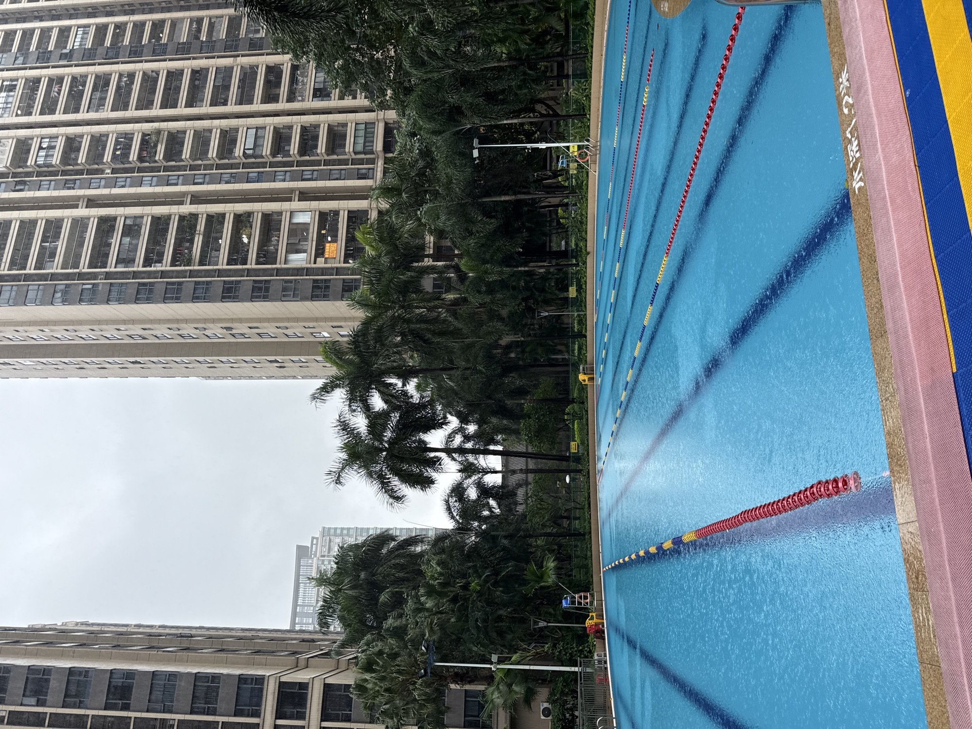 The image shows an outdoor swimming pool in Haizhu District, China, on a cloudy day. The pool is filled with clear blue water and has lane markers of yellow, blue, and red floating in it. Tall apartment buildings with many balconies loom in the background, along with lush palm trees and other greenery. There are a few yellow chairs visible around the edge of the pool, and a lifeguard chair with a red ring is also present. The water appears to be slightly rippled, indicating light rain or wind. A sign at the edge of the pool reads "水深1.2米," meaning "water depth 1.2 meters."