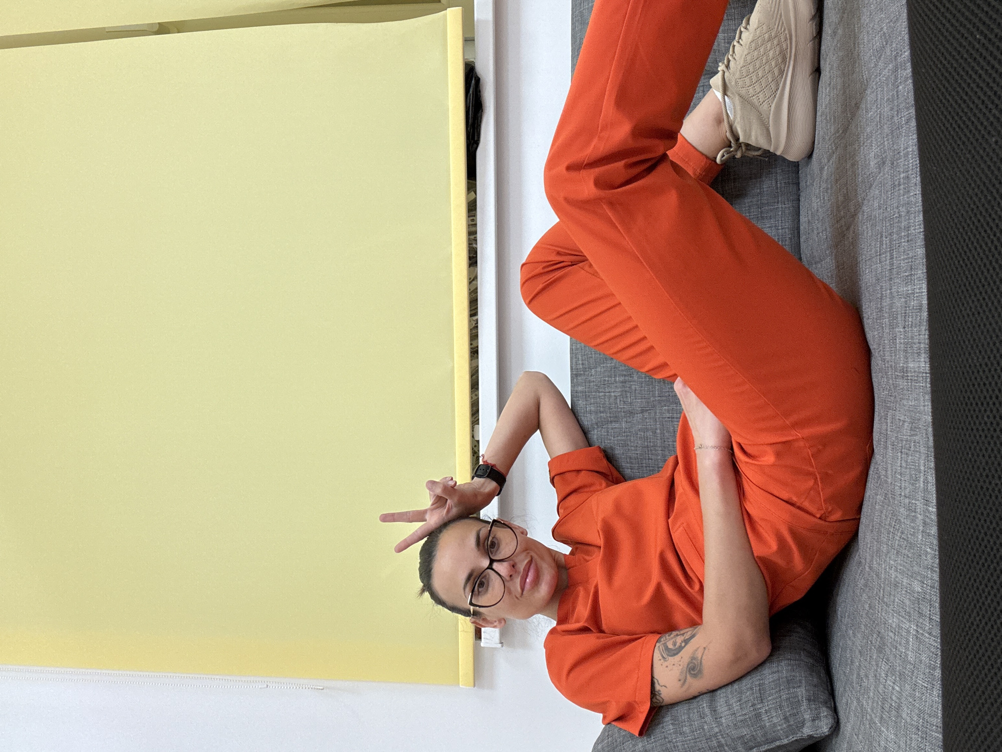 A woman wearing glasses and an orange jumpsuit is lying on a gray couch. She is holding up a peace sign with her left hand. She has a watch on her left wrist and tattoos on her left forearm. She is wearing tan sneakers. A yellow screen is visible in the background. The location is not discernible, and there is no evidence to suggest this image was taken in Pleven, Bulgaria.